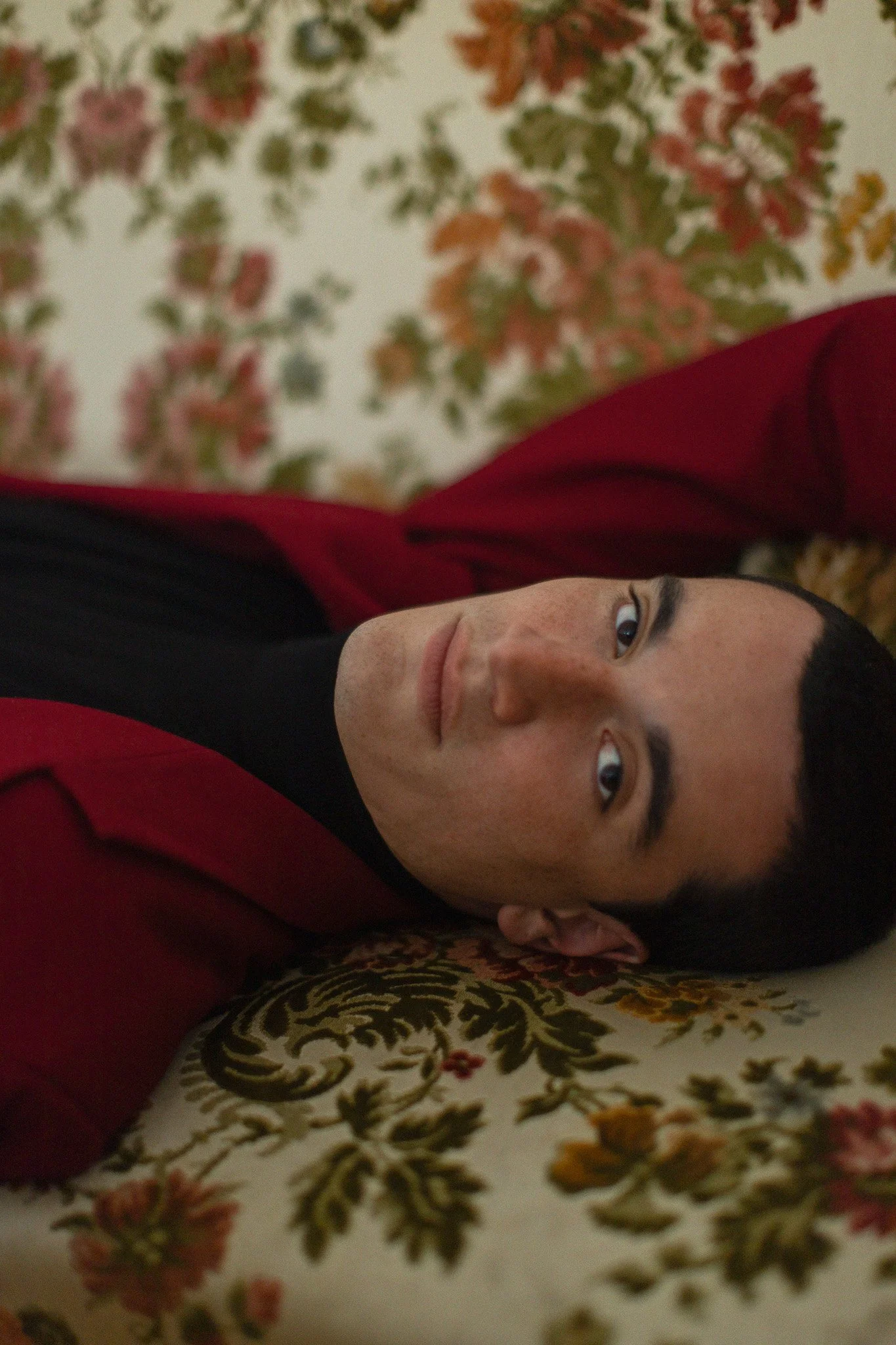 Jeune homme allongé sur un canapé à motifs floraux, regardant vers l'objectif, portant une veste rouge et un haut noir.