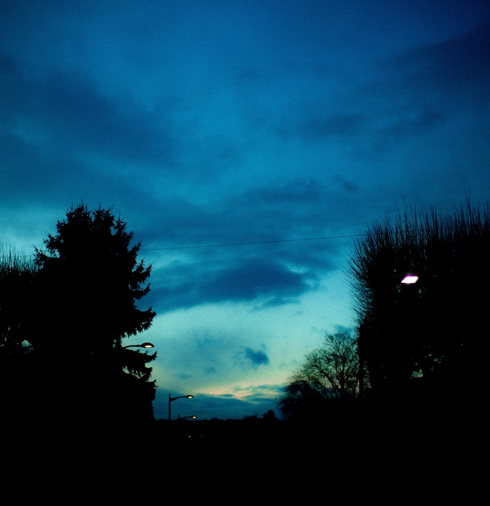 Ciel crépusculaire avec nuages, silhouettes d'arbres et lampadaires.