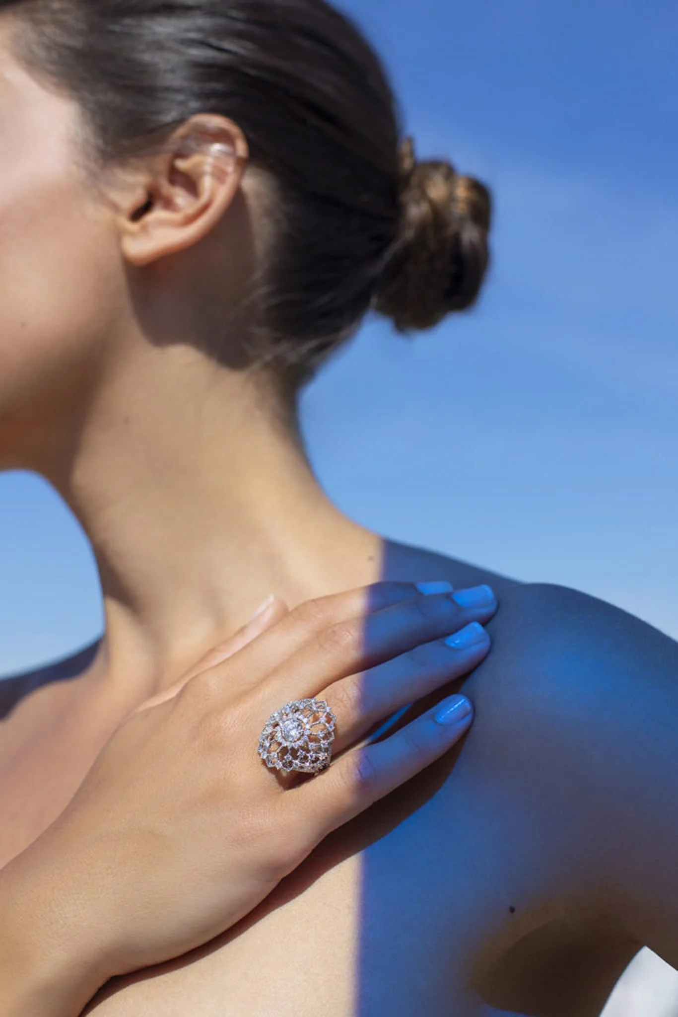 Femme avec cheveux bruns en chignon, portant un anneau de diamant en forme de fleur, main sur l'épaule, ciel bleu en arrière-plan.