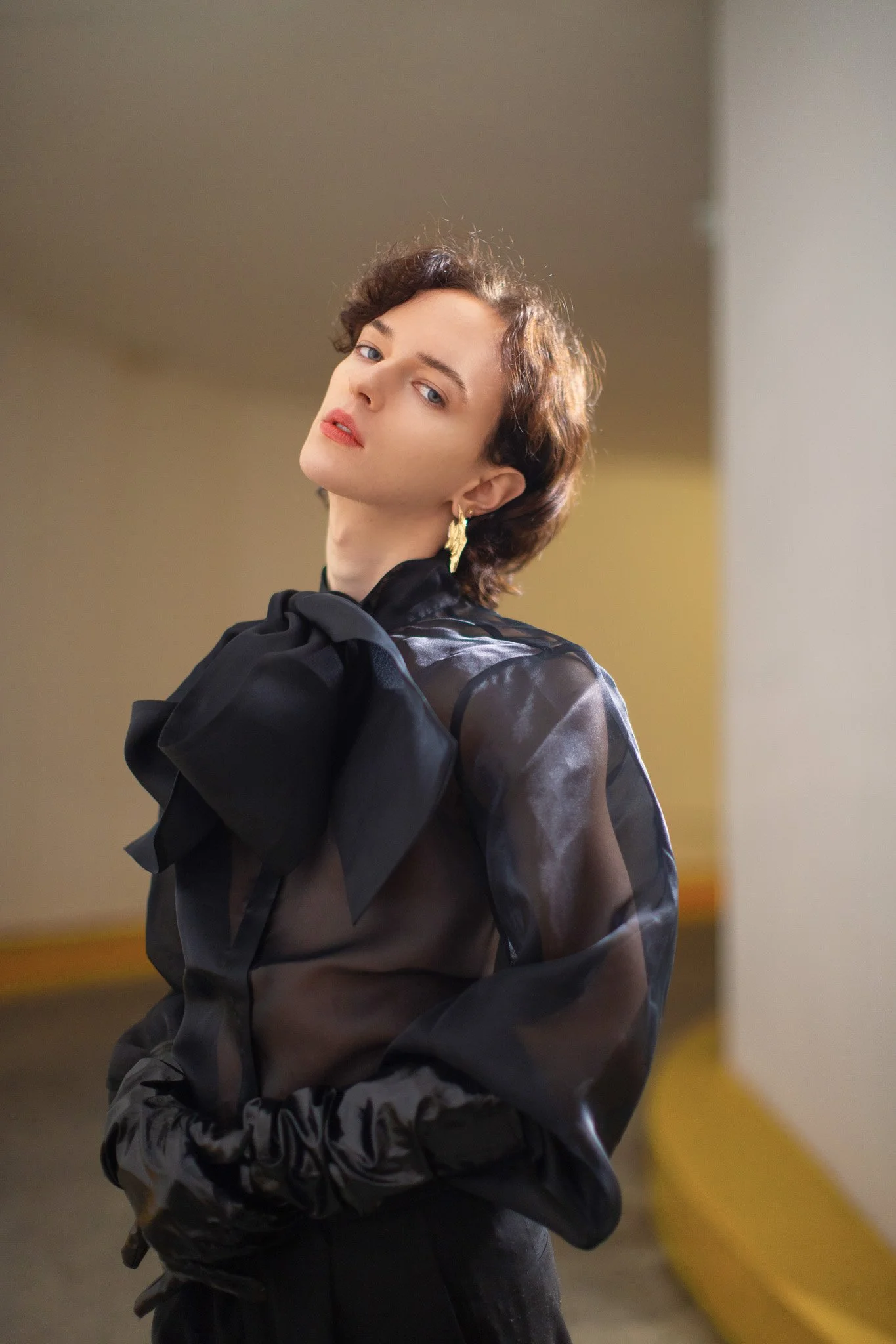 Femme avec des cheveux courts, portant une blouse en tissus transparent noir avec un grand nœud sur l'épaule, portant des boucles d'oreilles dorées, dans un environnement intérieur, regardant vers l'objectif.