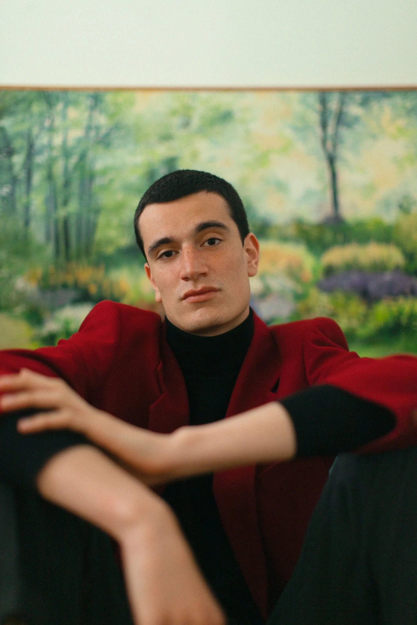 Un jeune homme en veste rouge assis avec une personne dont on voit seulement un bras, avec un fond de paysage nature en peinture.