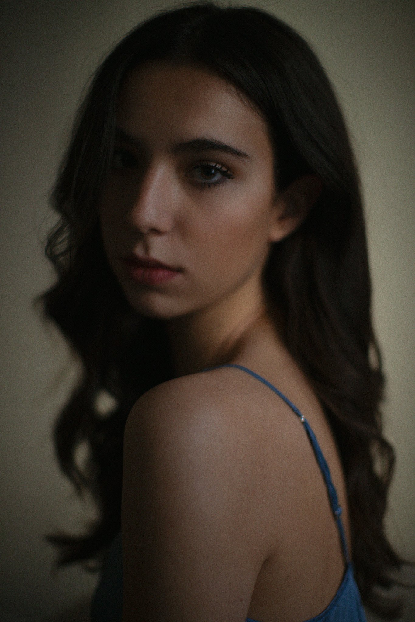 Portrait d'une jeune femme avec de longs cheveux bruns bouclés, portant une robe bleue à bretelles fine, regardant légèrement vers la caméra dans une lumière douce.