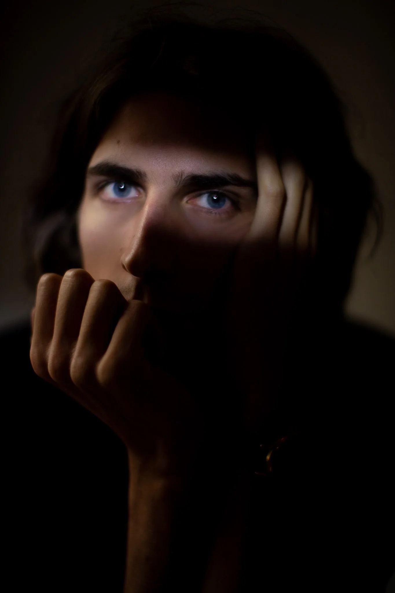 Portrait sombre d'une personne aux yeux bleus, tenant sa tête avec une main, dans un éclairage faible.