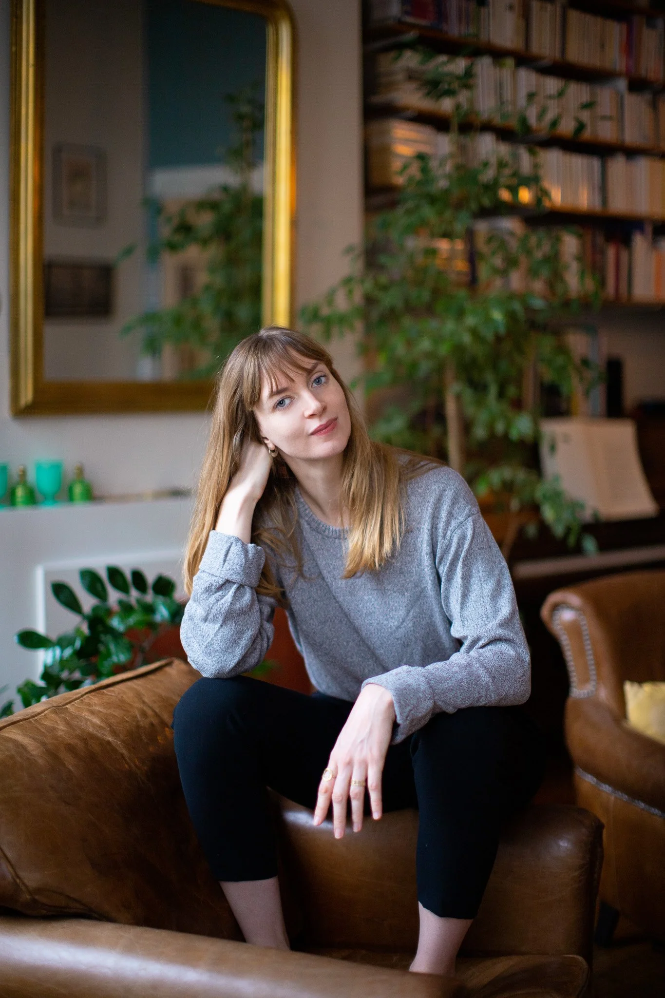 Une femme aux cheveux châtains clairs et aux yeux bleus, assise sur un canapé en cuir marron, dans un salon avec des étagères remplies de livres, une grande plante verte, et un miroir doré en arrière-plan.
