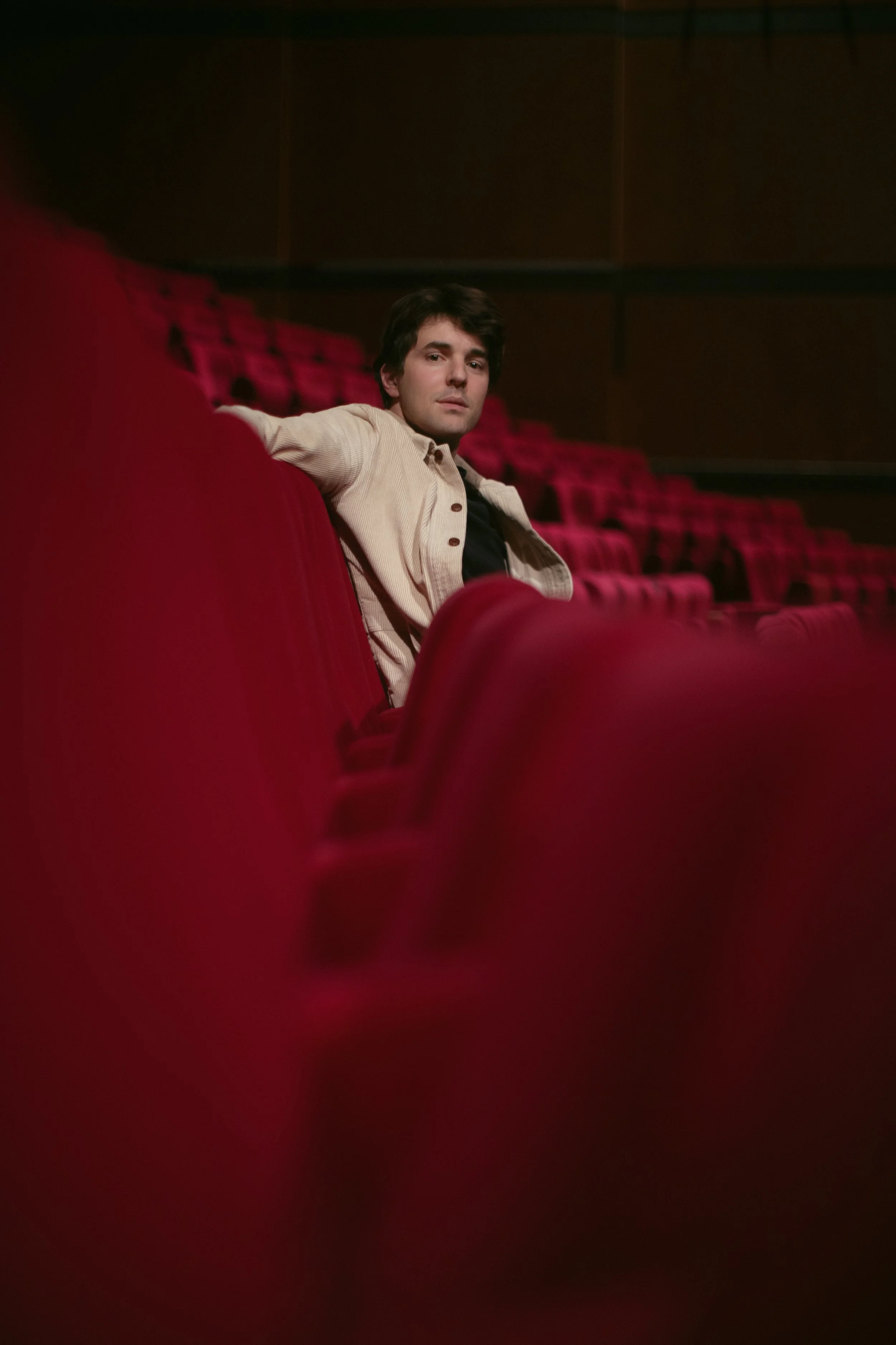 Un jeune homme assis dans une salle de cinéma vide avec des sièges rouges, regardant vers la caméra.