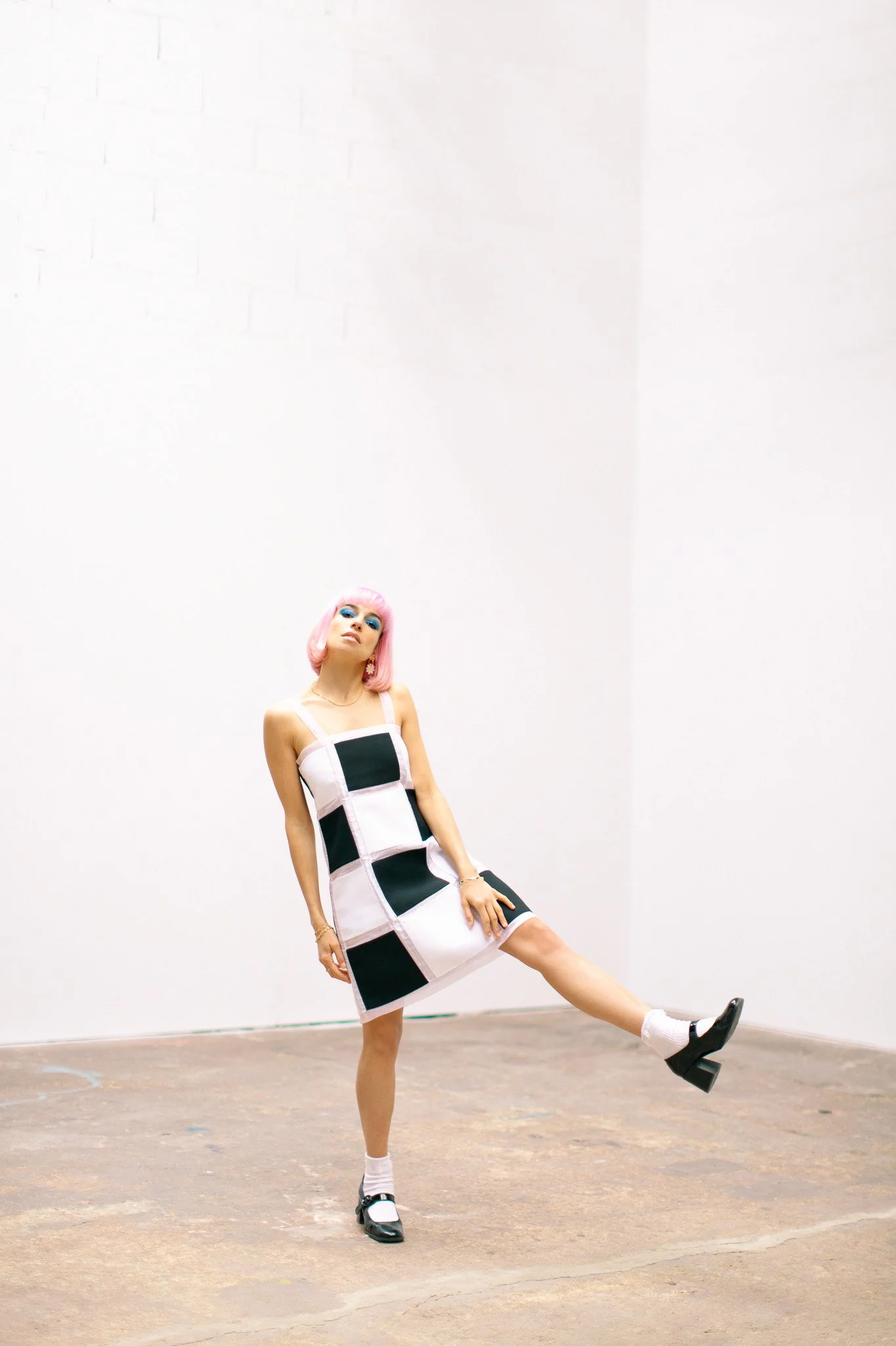 Une femme avec une perruque rose, portant une robe blanche et noire à motifs géométriques, se tient dans une pièce aux murs blancs, posant avec une jambe levée et un air confiant.
