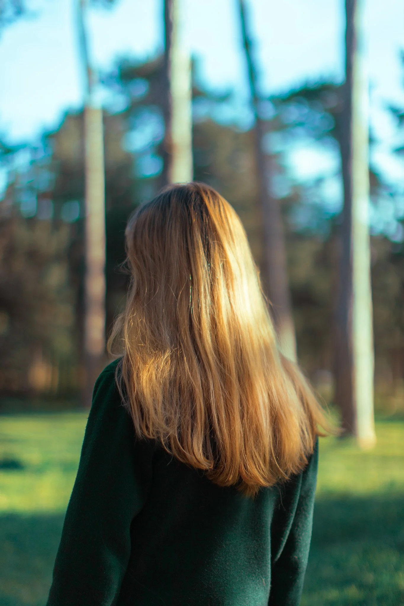 Une femme aux cheveux longs et blonds vue de dos, portant un pull vert, dans un parc avec des arbres et un ciel bleu.