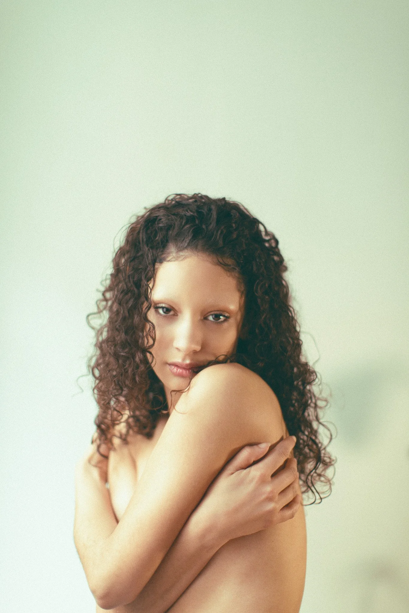 Une femme avec des cheveux bouclés qui se couvre avec ses bras, regardant l'objectif avec une expression douce, fond blanc