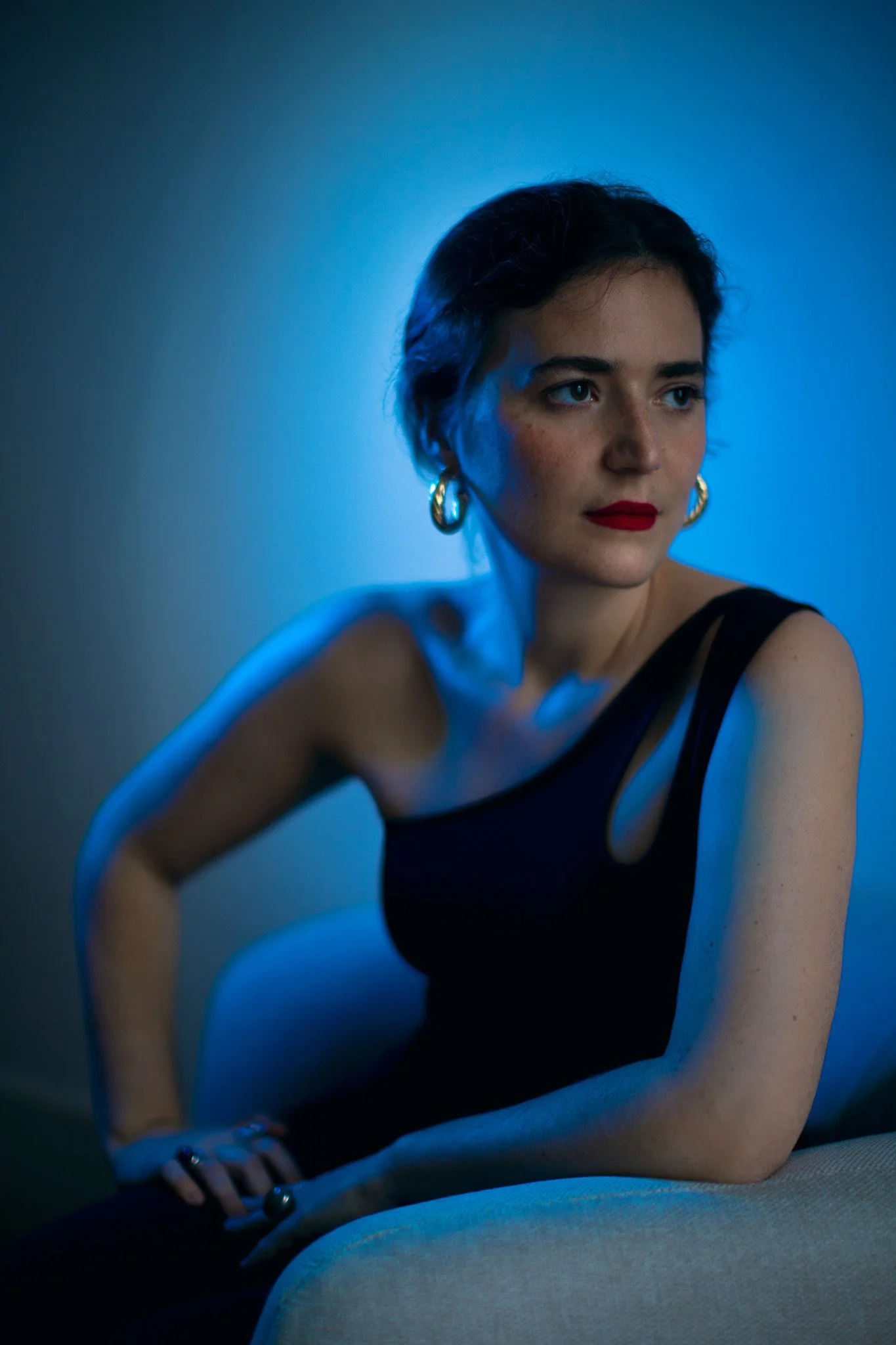 Une femme avec des cheveux courts, portant un haut noir, assise sur un canapé beige, dans un éclairage bleu, regardant vers la droite.
