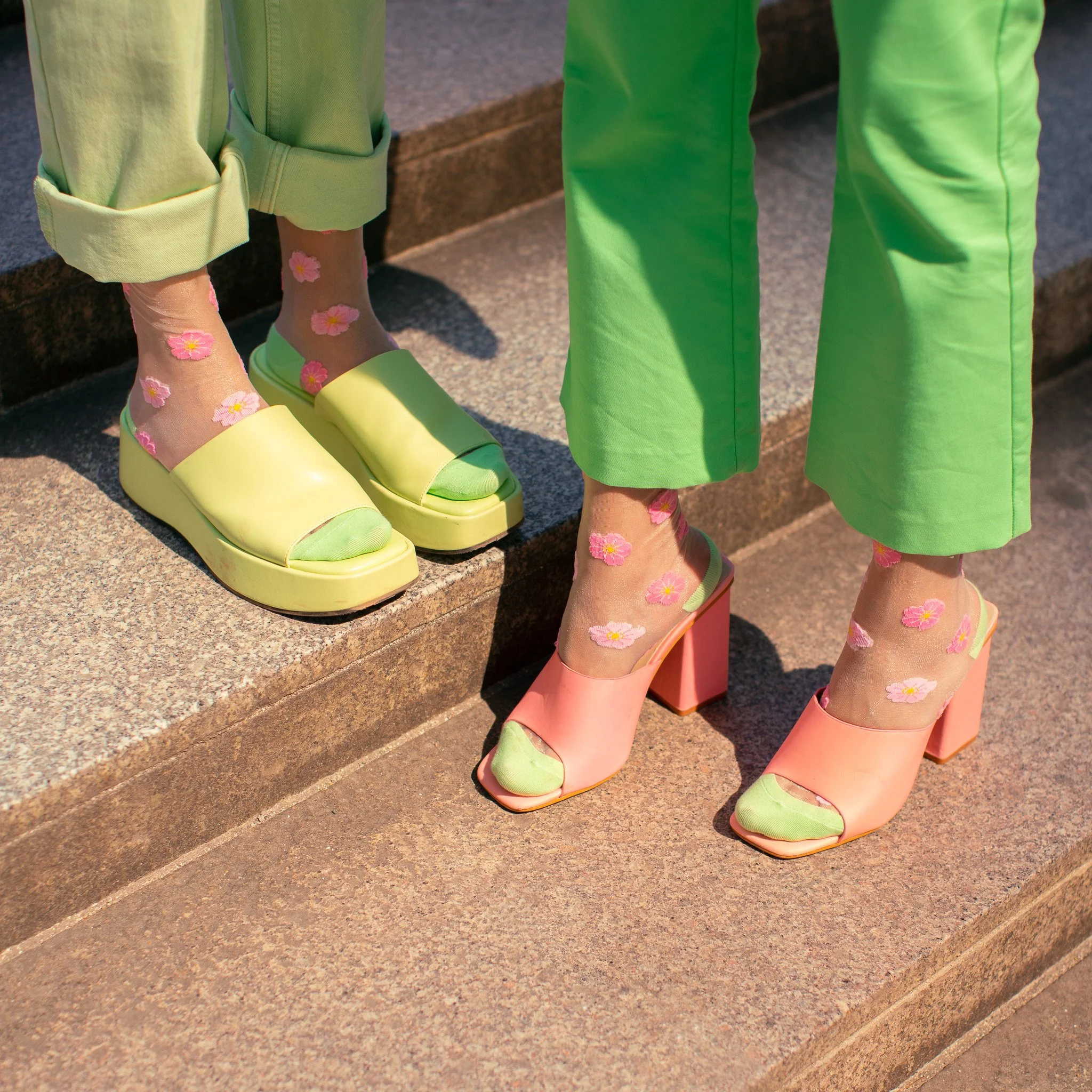Deux personnes portent des chaussures à talons hauts, accompagnées de chaussettes transparentes avec des motifs de fleurs roses et jaunes, sur des marches en pierre.