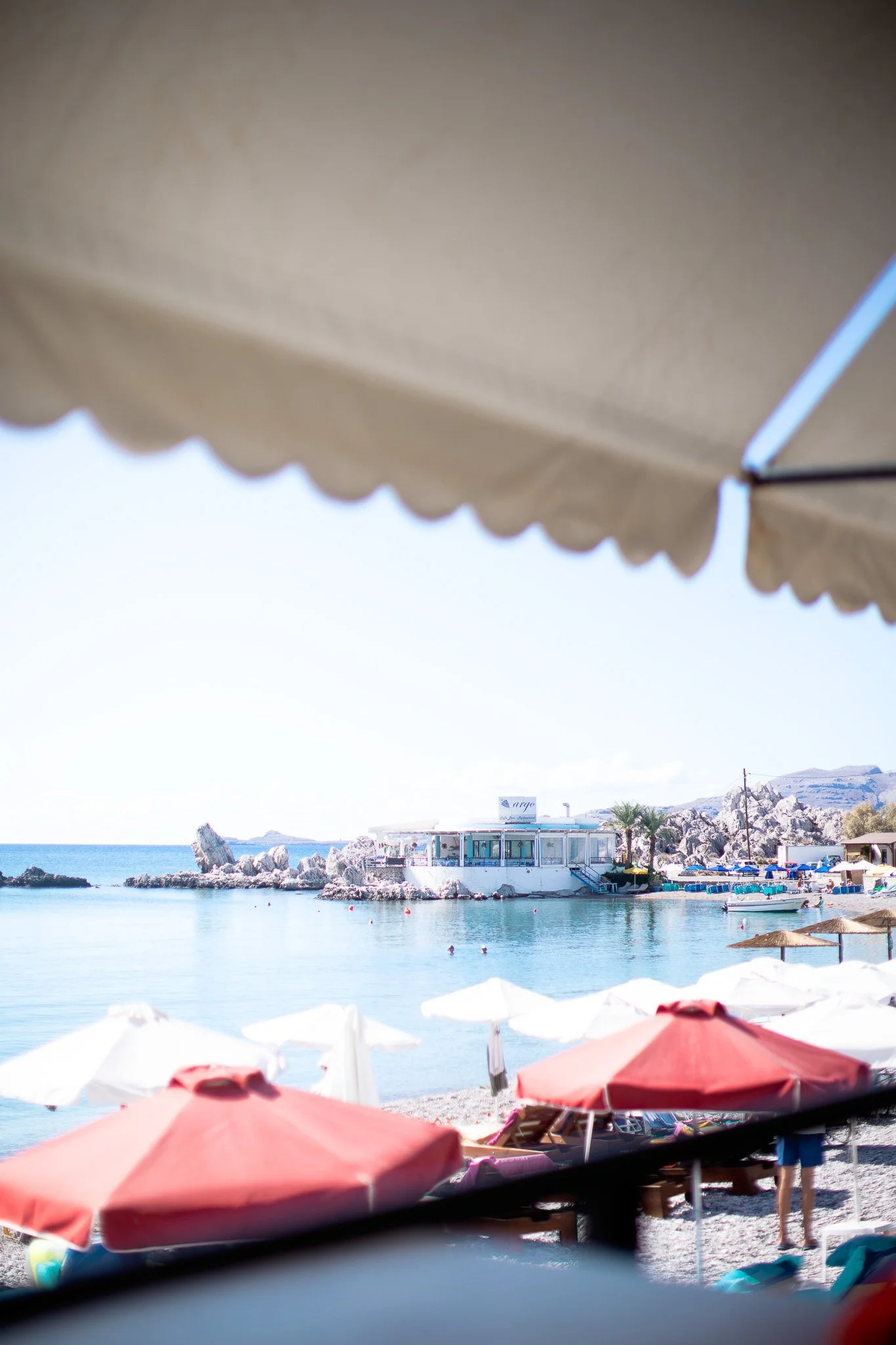 Plage avec parasols rouges et blancs, eau claire, bateau, bâtiment en arrière-plan, rochers, montages et palmier.