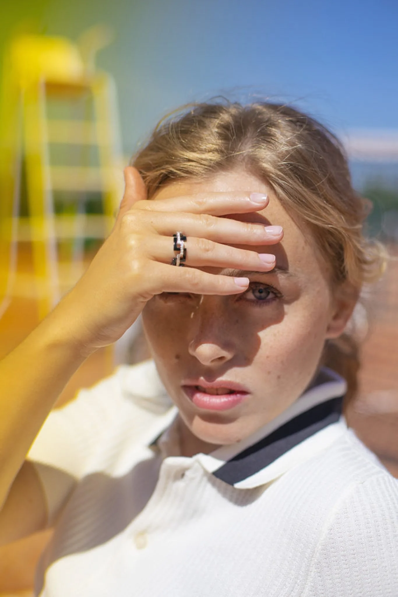 Jeune femme avec cheveux courts, portant une chemise blanche avec col rayé, tenant sa main sur son visage avec un doigt sur son front, dans un espace en plein air sous un ciel bleu.