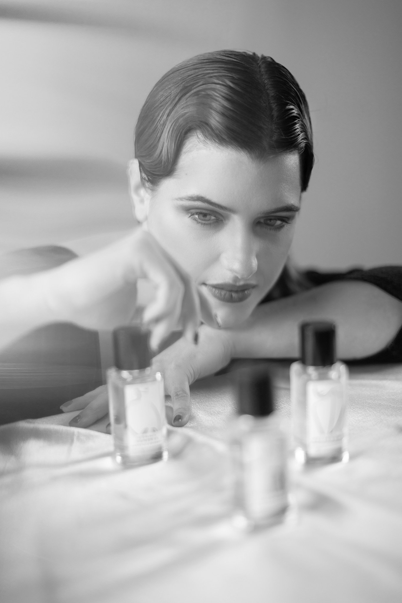 Une femme aux cheveux courts, en noir et blanc, regarde fixement vers le haut, entourée de flacons de vernis à ongles sur une surface