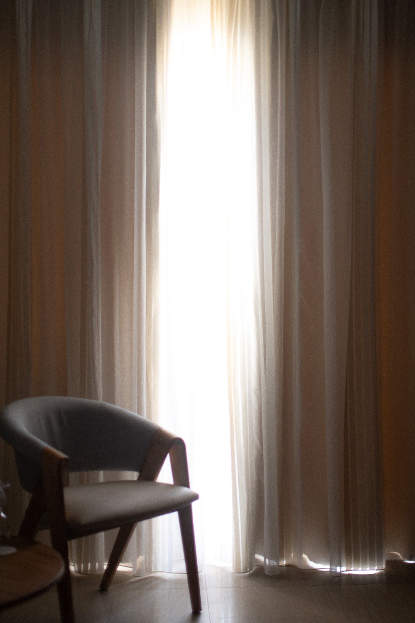 Une fenêtre avec des rideaux blancs lumineux, une chaise en bois avec un coussin attrape-soleil, dans une pièce calme.