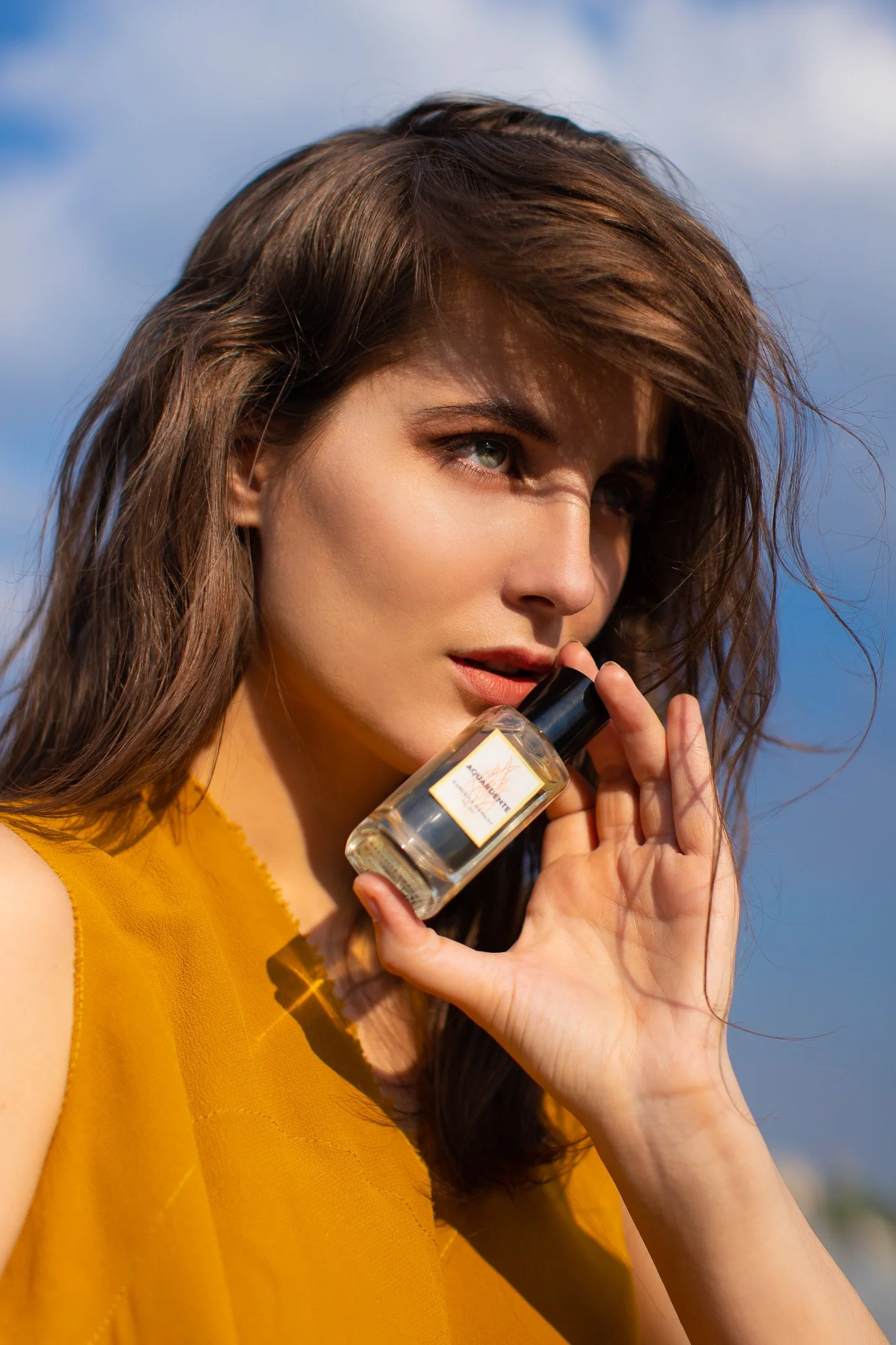 Jeune femme avec des cheveux bruns, portant une robe jaune, tenant un flacon de parfum, en plein air sous un ciel partiellement nuageux.