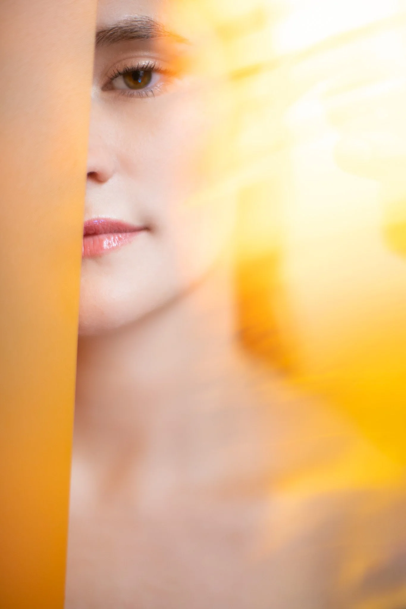 Un gros plan du visage d'une femme avec un fond lumineux jaune et orange, moitié cachée derrière une surface orange.