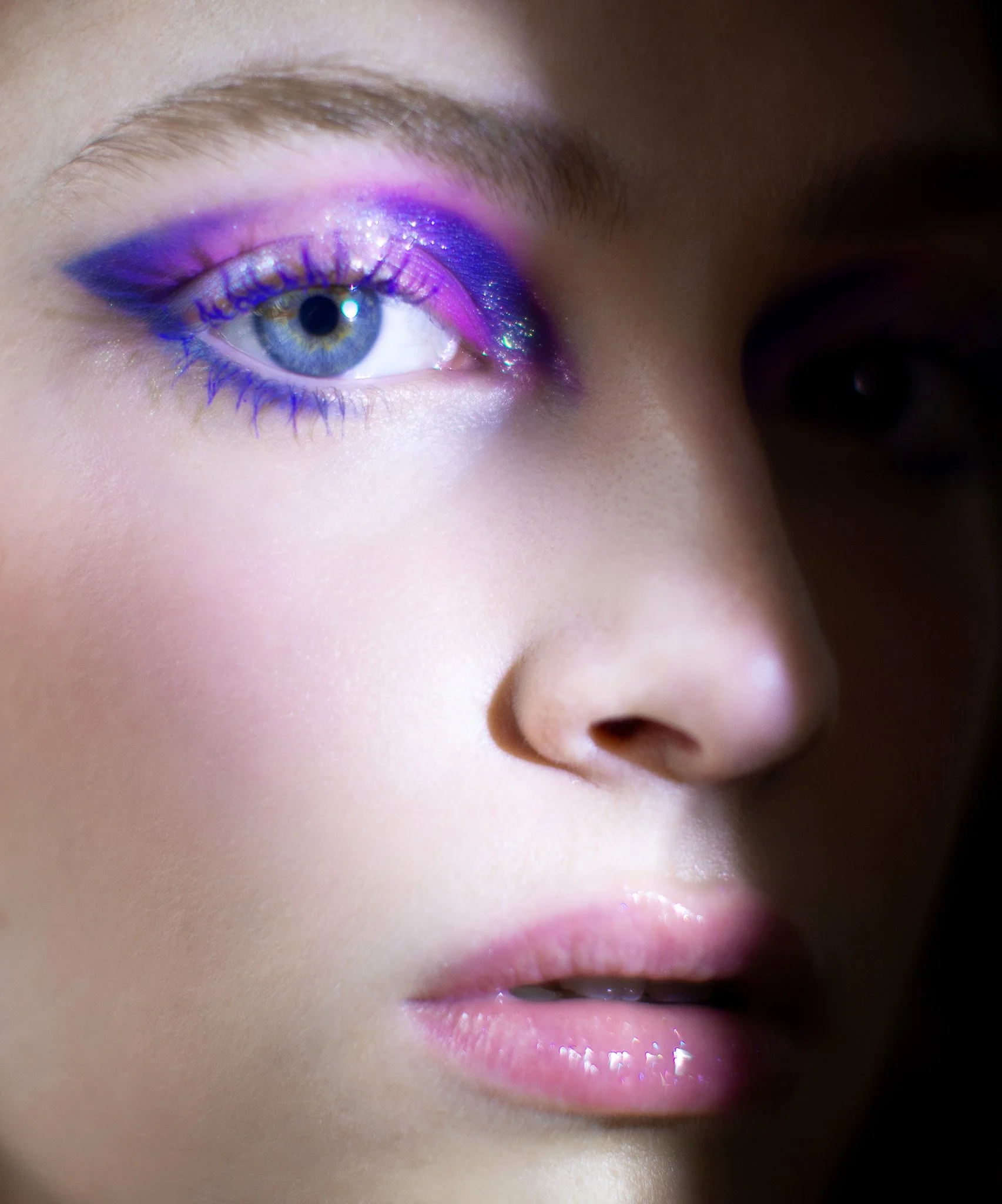 Femme avec maquillage coloré violet, rose et bleue, portant du maquillage brillant autour de l'œil, visage en gros plan.