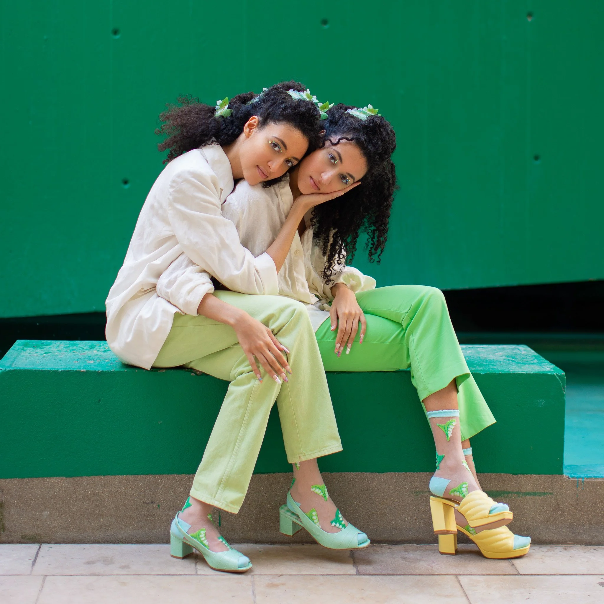 Deux femmes assises l'une contre l'autre sur un banc, portant des vêtements clairs et des chaussures à talons colorés, avec un fond vert.