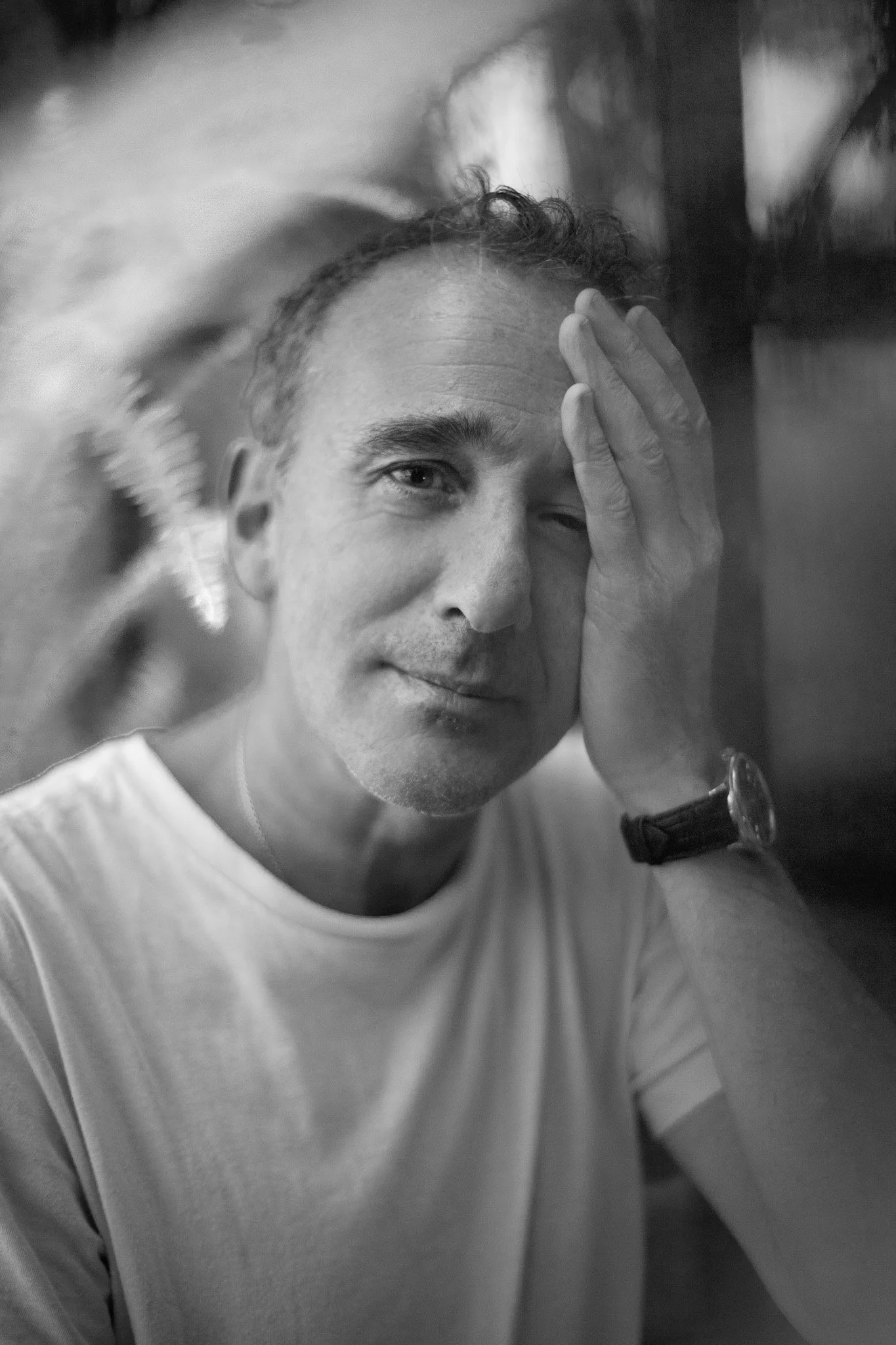 Homme en t-shirt blanc, portant une montre, posé contre un mur, faisant un clin d'œil et couvrant une partie de son visage avec sa main.