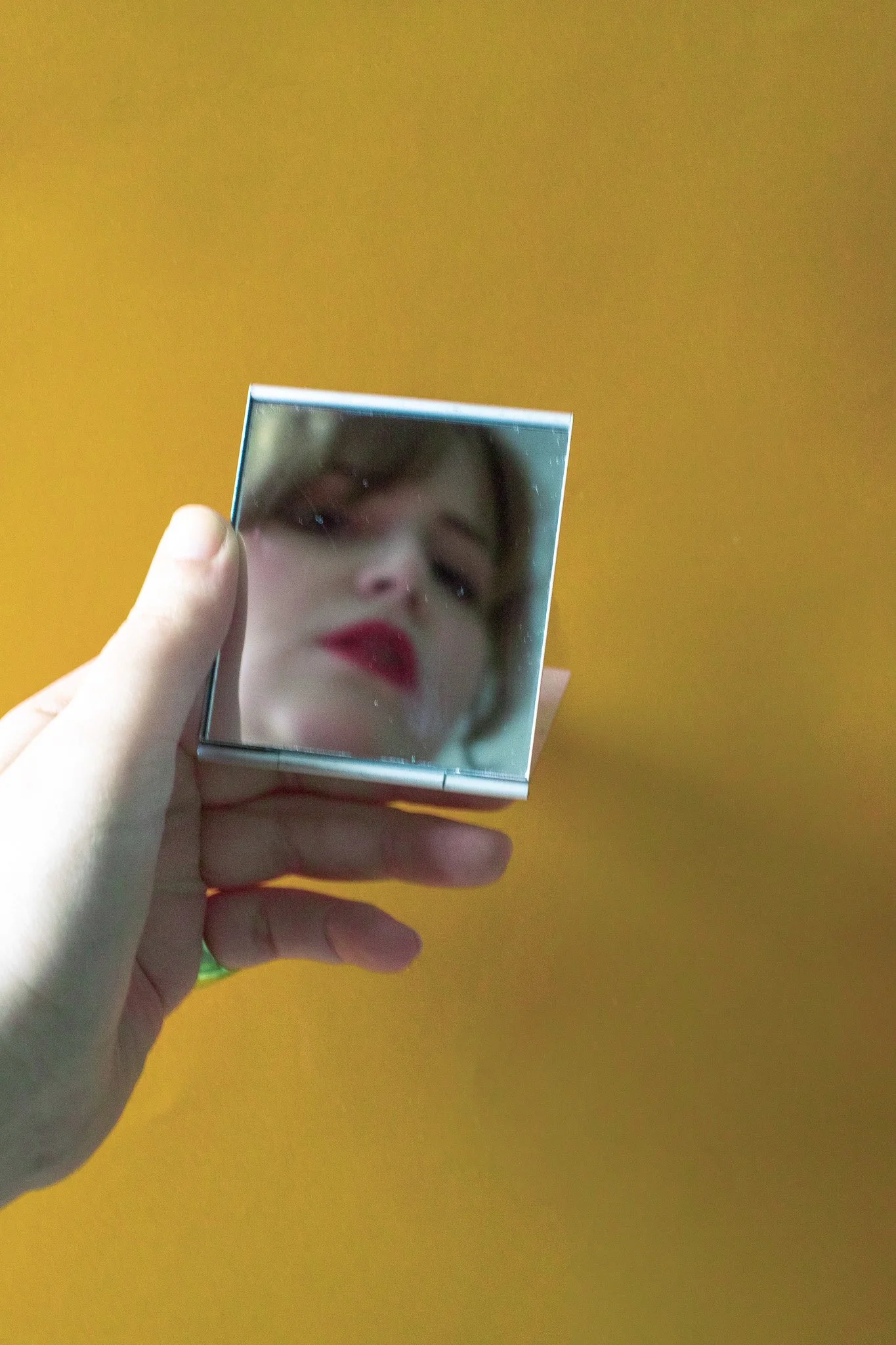 Une main tient un miroir de poche réfléchissant le visage d'une femme portant du rouge à lèvres rouge, avec un fond de couleur jaune-orange.