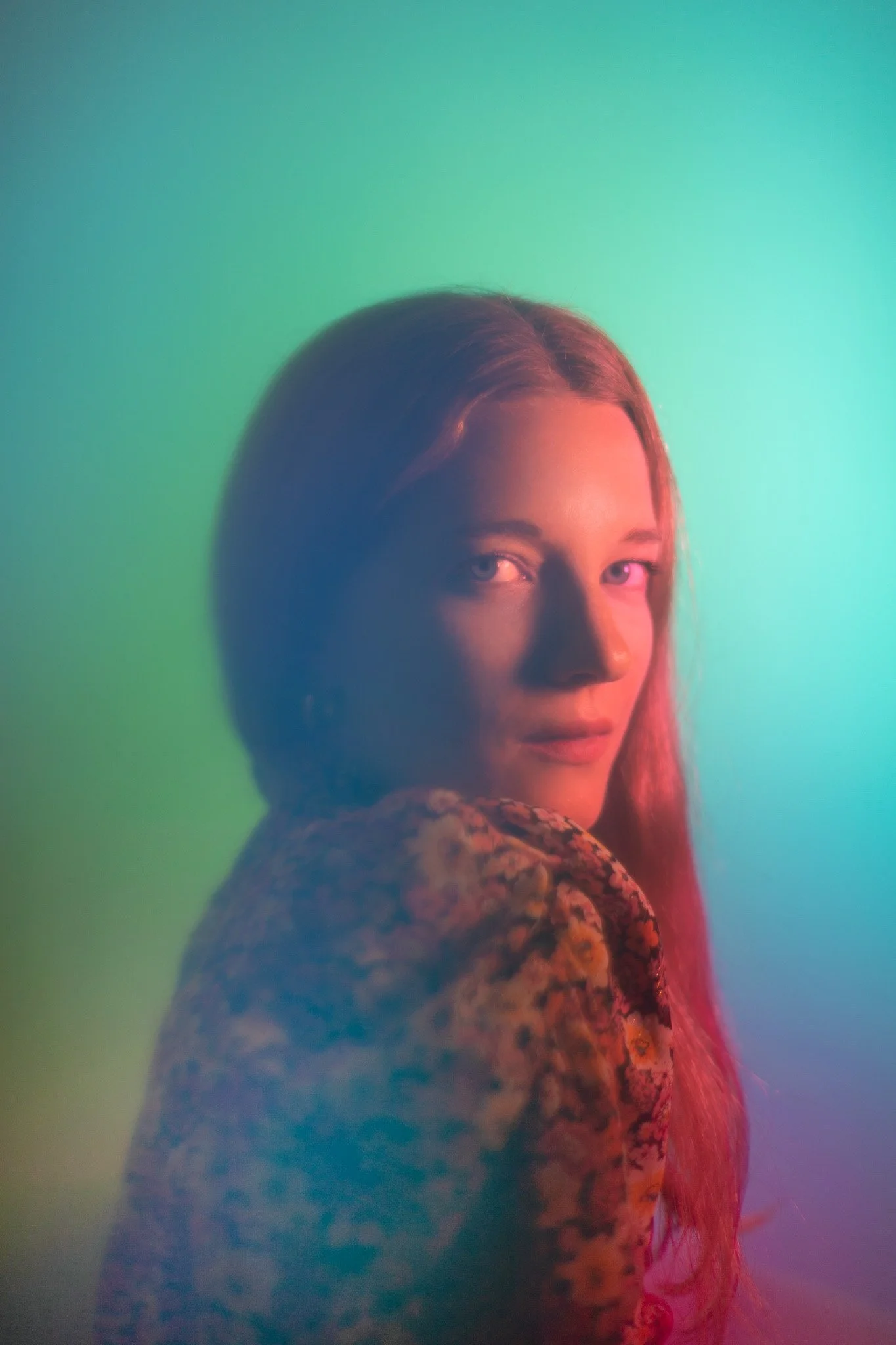 Portrait d'une jeune femme aux cheveux longs, regardant par-dessus son épaule, avec un fond coloré dégradé en vert et bleu.