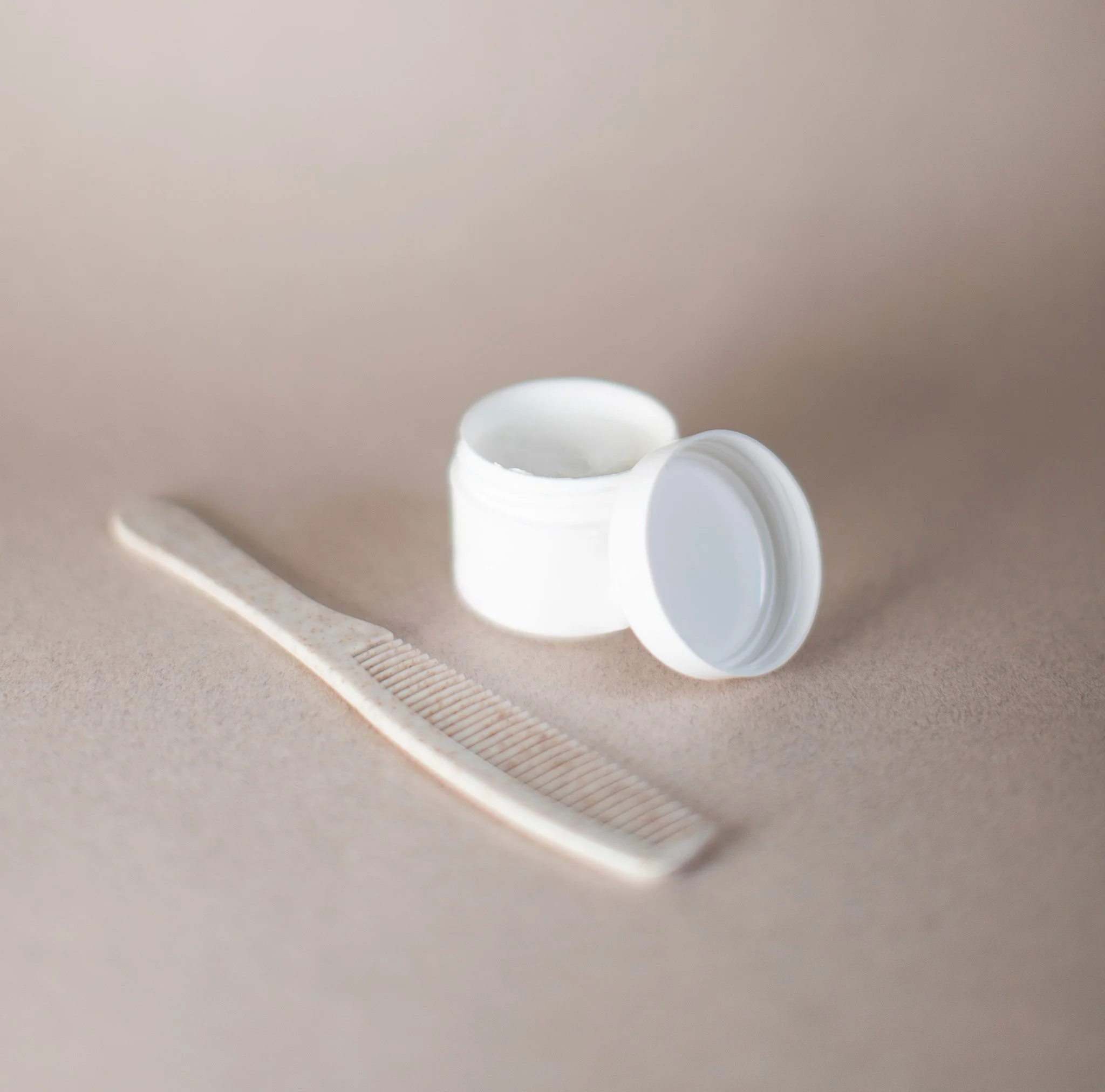 Un pot blanc de crème ou de gel, ouvert, et un peigne blanc en plastique sur un fond beige clair.