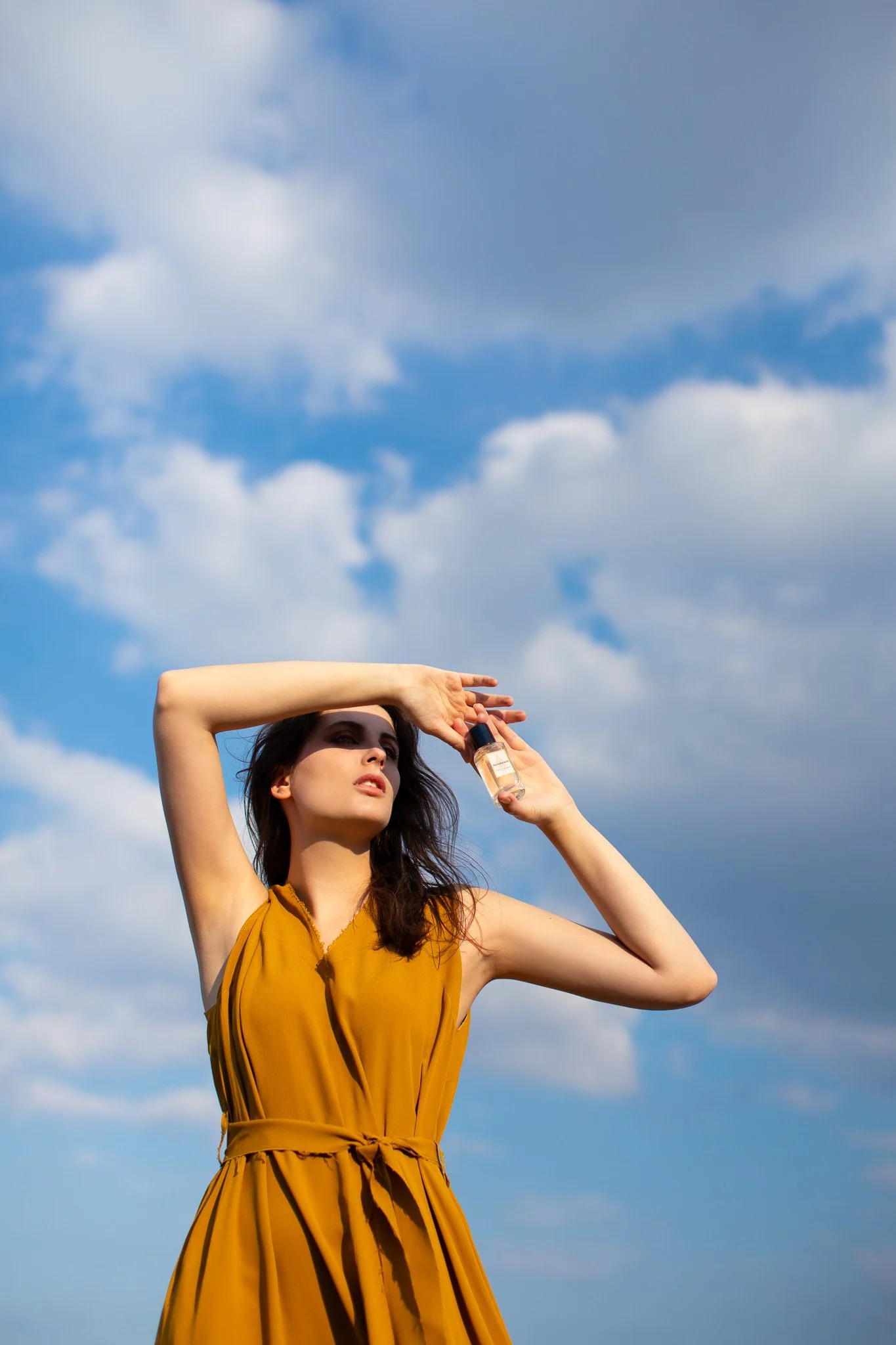 Une jeune femme en robe jaune tient un flacon de parfum en se protégeant du soleil avec sa main contre un ciel bleu avec des nuages blancs.