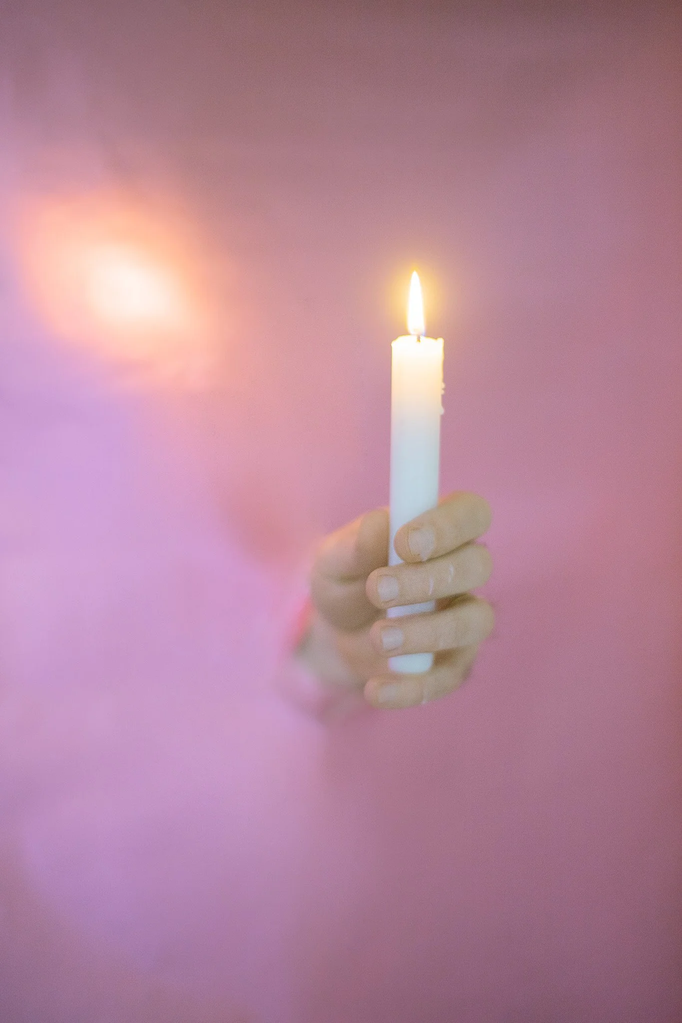 Main tenant un bougie blanche allumée contre un fond rose, avec un reflet de lumière sur la surface rose.