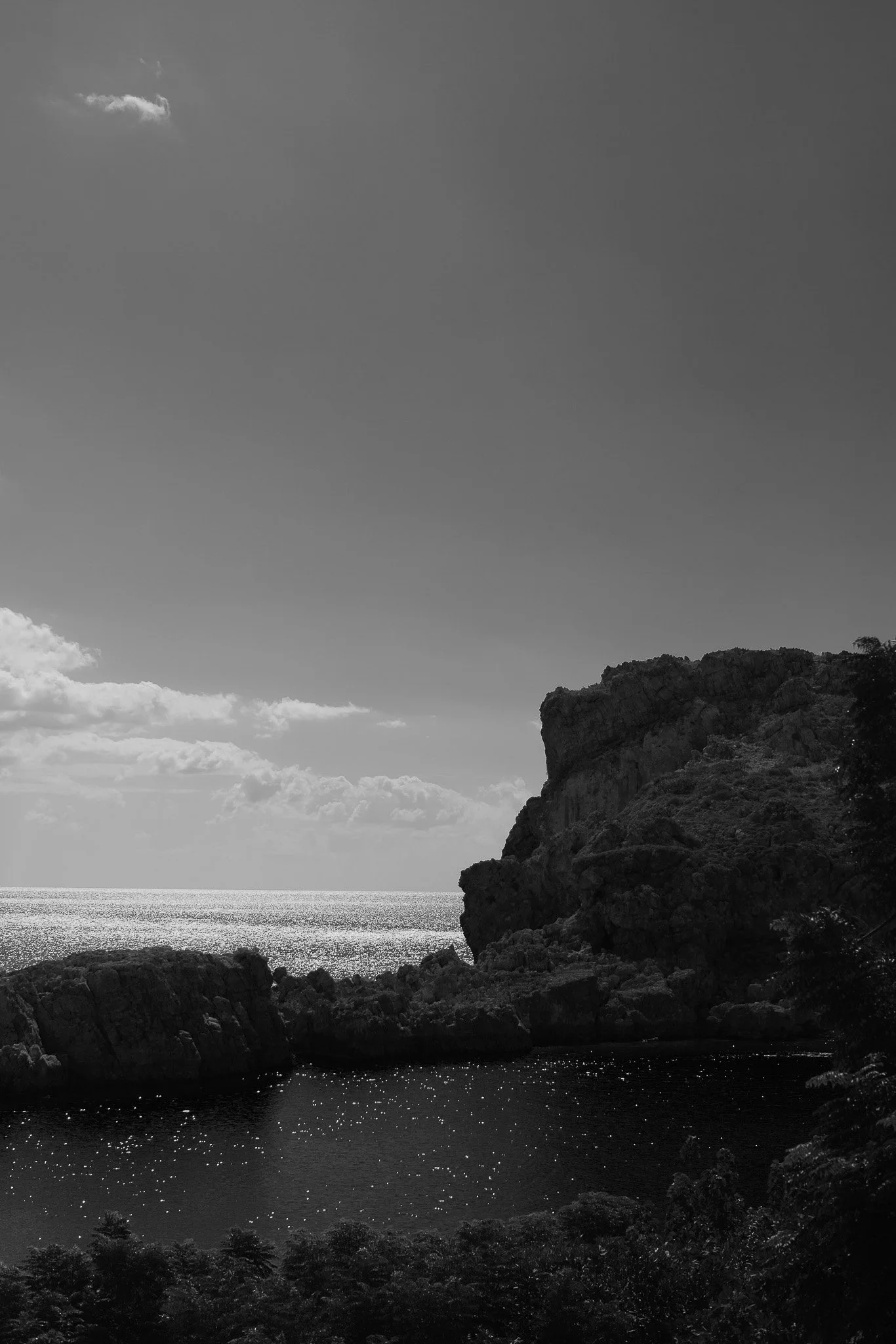 Cliché en noir et blanc d'une côte rocheuse avec la mer en arrière-plan, quelques nuages dans le ciel et les reflets du soleil sur l'eau.