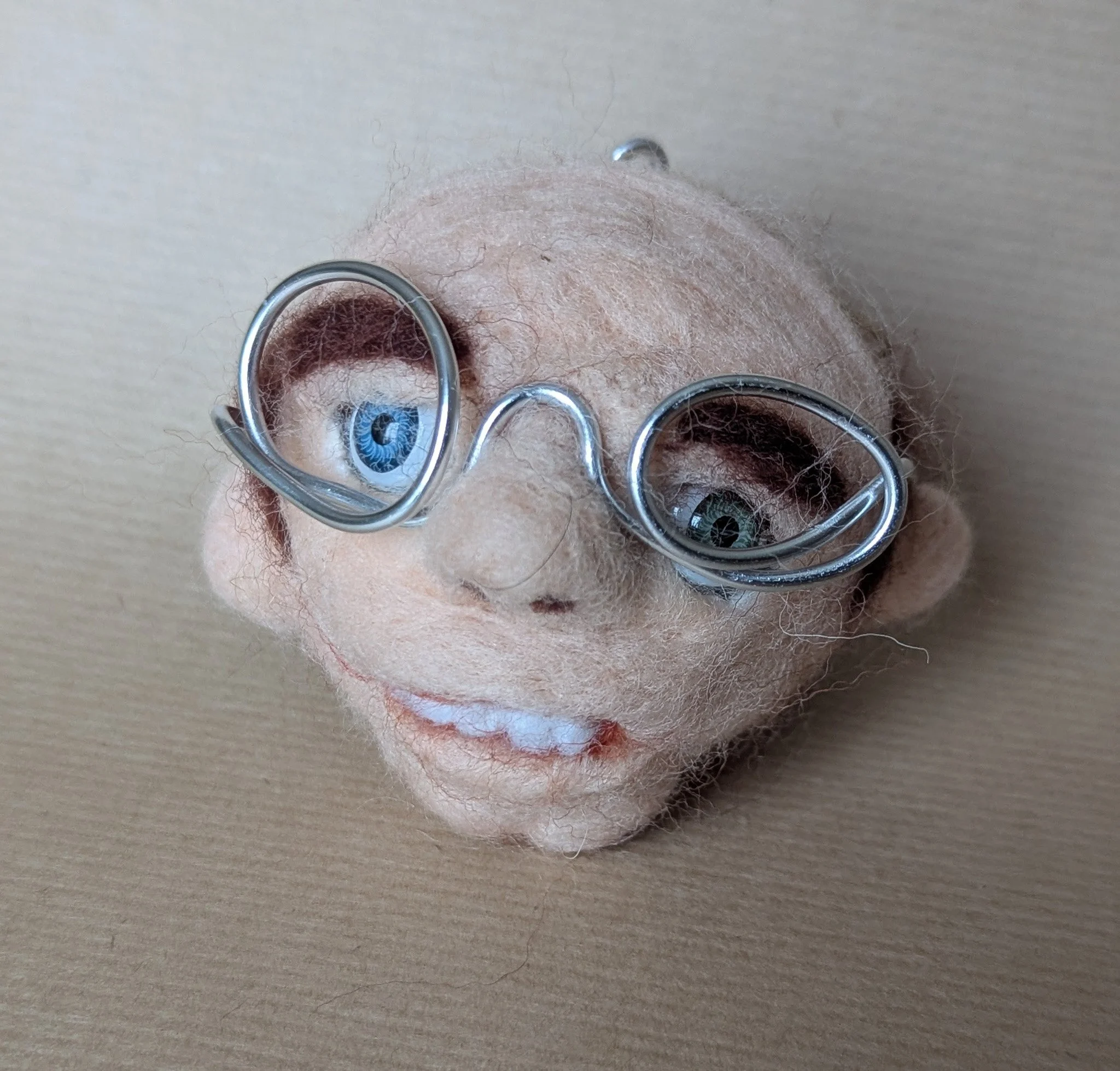 Tête de poupée enfeutrée avec des lunettes amusantes en fil métallique, ayant des yeux bleus et marron, sur une surface beige.
