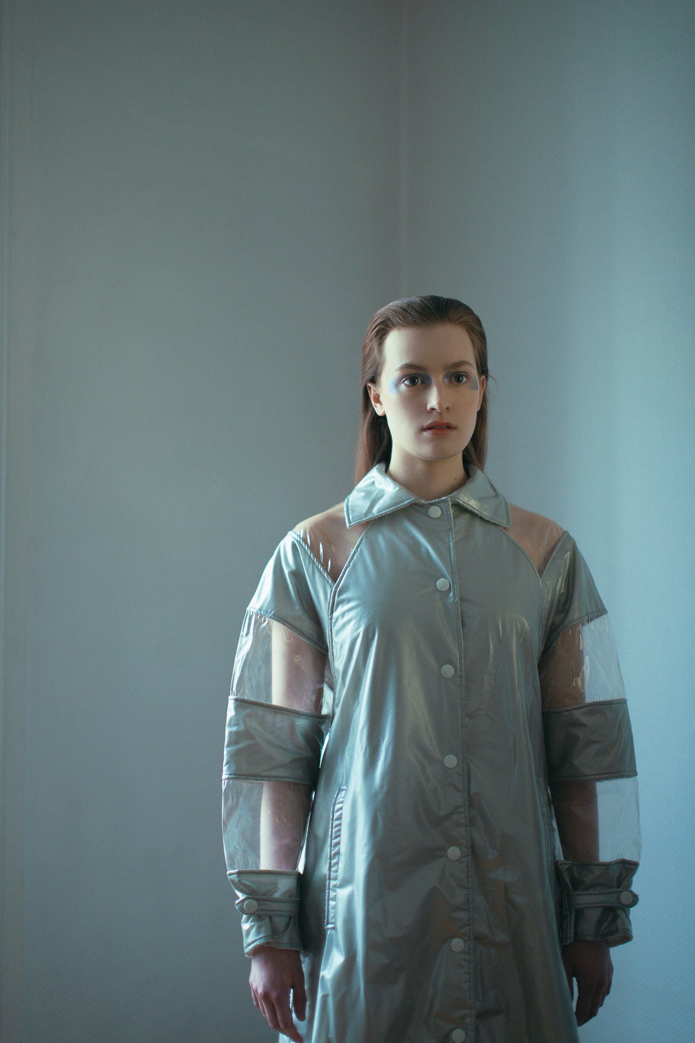 Une femme avec des cheveux bruns, porte une veste en matériau brillant argenté et transparent, debout dans une pièce avec un mur gris clair en arrière-plan.