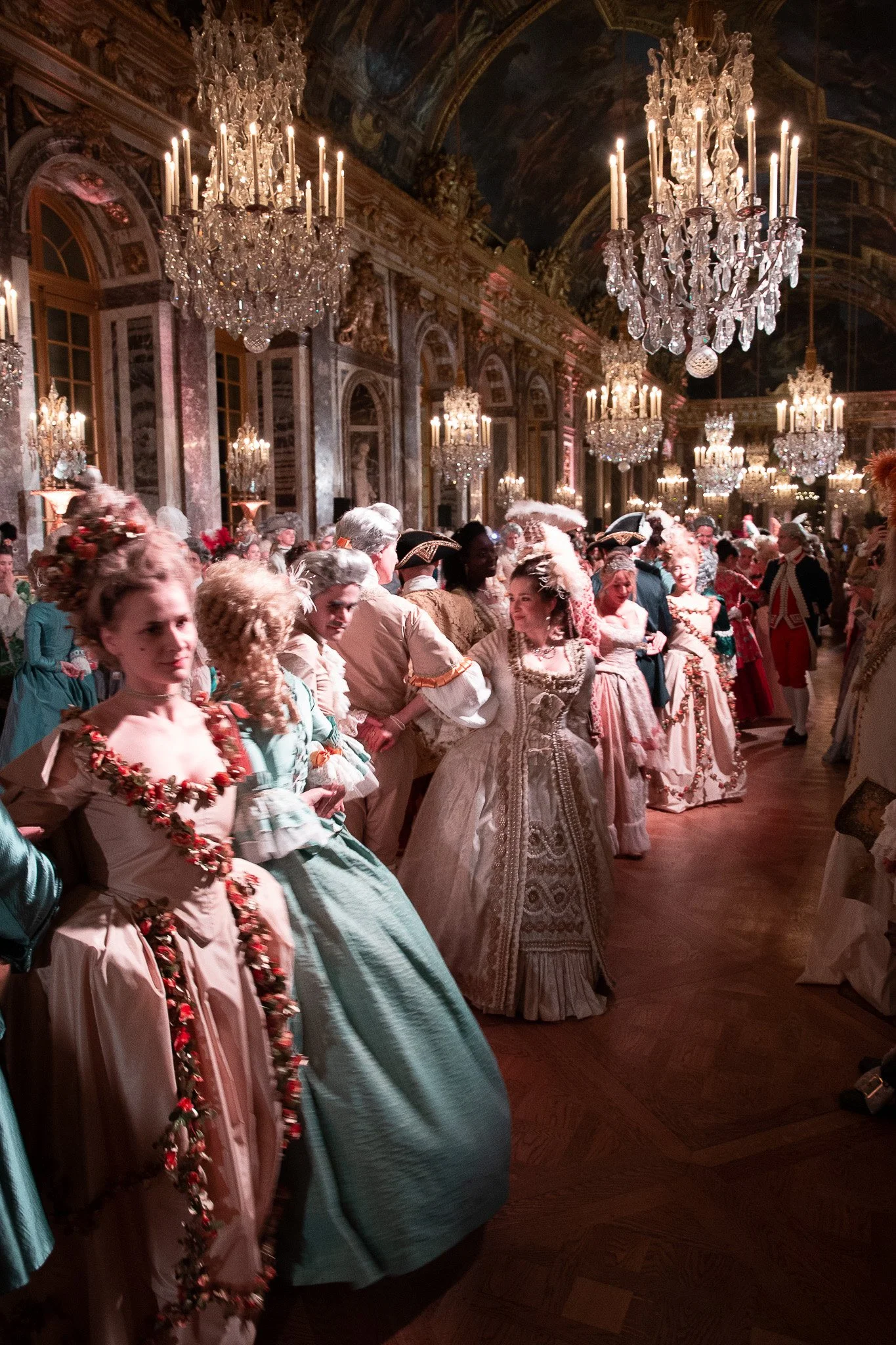 Registre de personnes portant des vêtements historiques dans une salle ornée de chandeliers en cristal, avec un plafond peint et des murs décorés dans un style baroque.