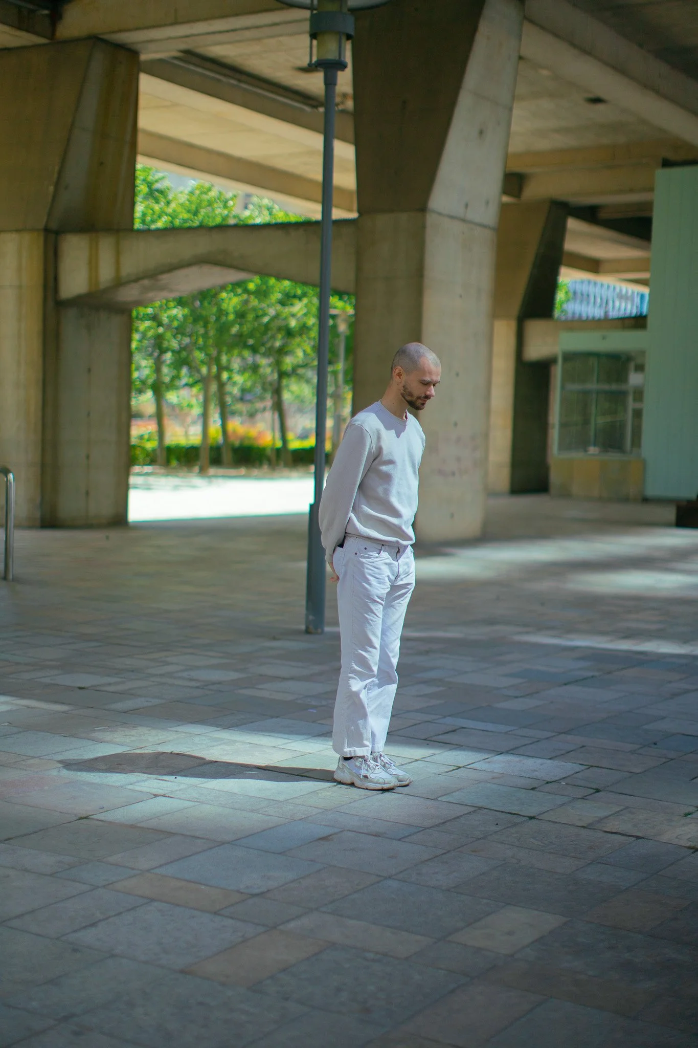 Un homme en vêtements blancs se tient seul sur un sol pavé sous un pont ou une structure en béton, avec des arbres verts en arrière-plan.