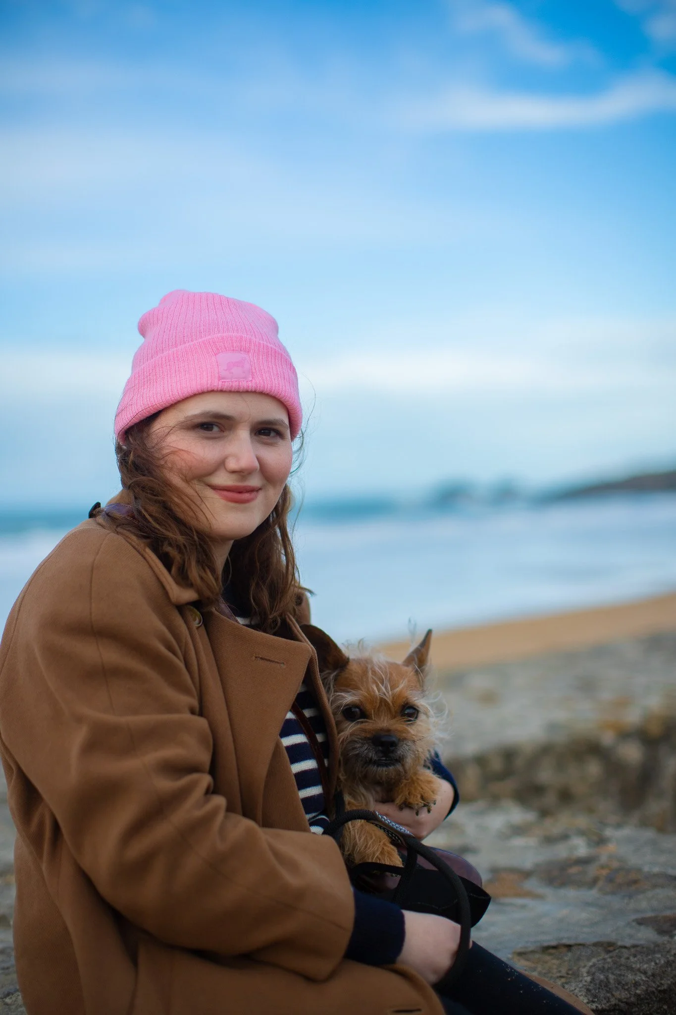 Jeune femme souriante portant un manteau marron, un bonnet rose, assise sur la plage, tenant un petit chien brun.