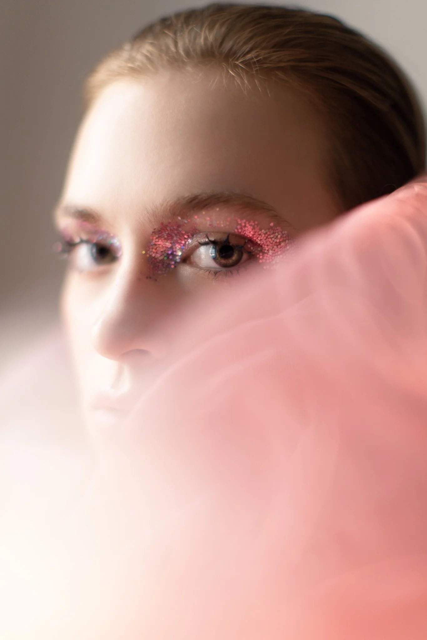 Un portrait d'une jeune femme avec un maquillage brillant, notamment autour des yeux avec des paillettes roses et multicolores, et une étreinte douce en rose dans le premier plan.