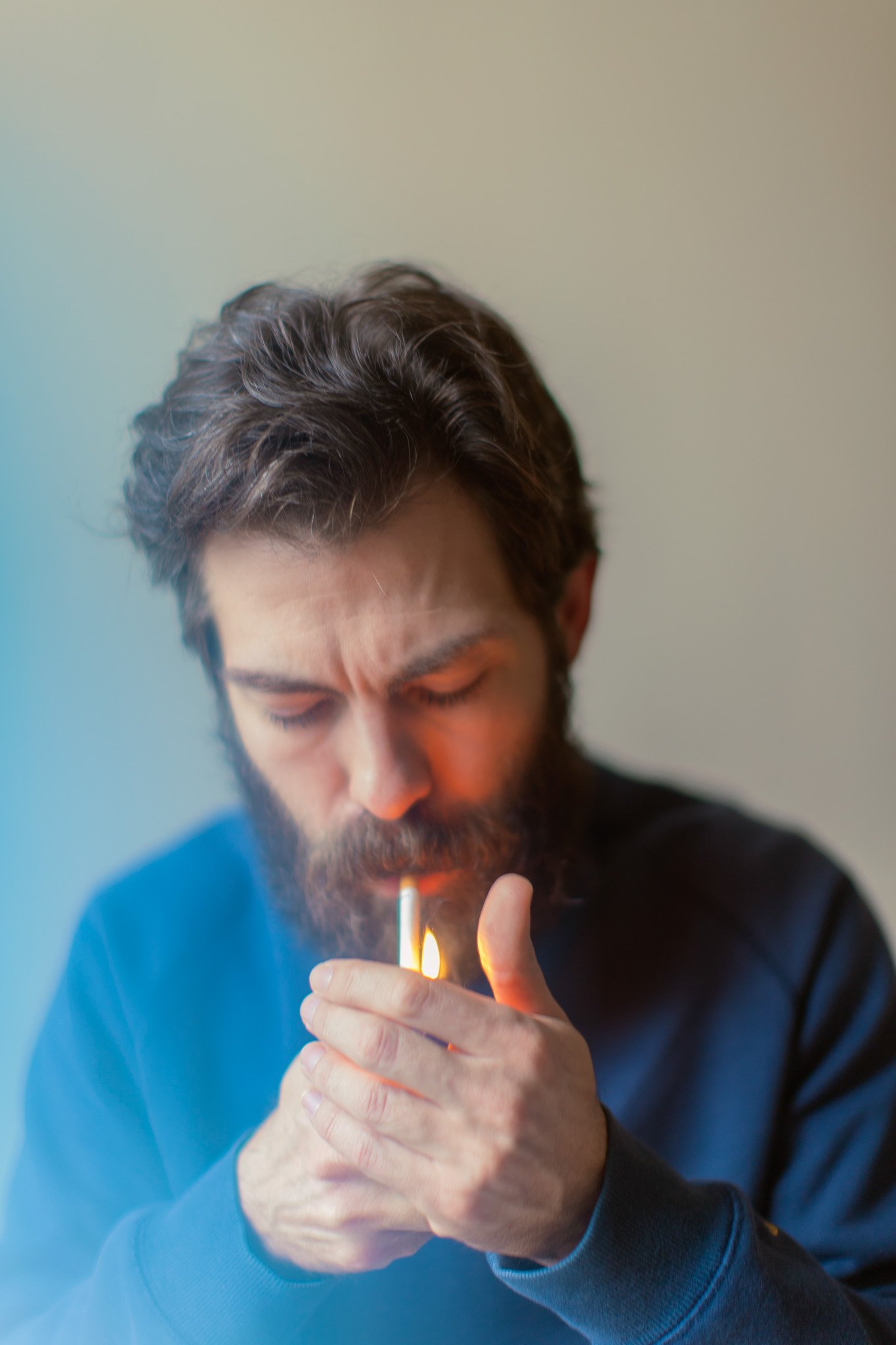 Homme avec une barbe, concentré, allumant une cigarette avec un briquet flame, portant un sweat noir, fond neutre.