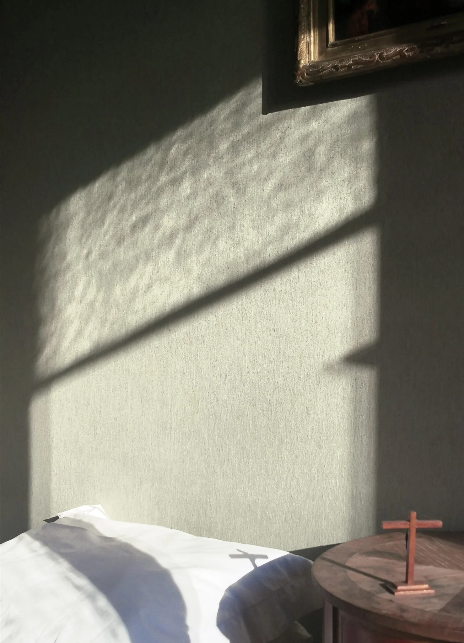 Une table ronde en bois avec une petite croix sur le dessus, une partie d'un lit avec une couverture blanche, et un mur gris avec des ombres de rayures et un cadre en or suspendu.