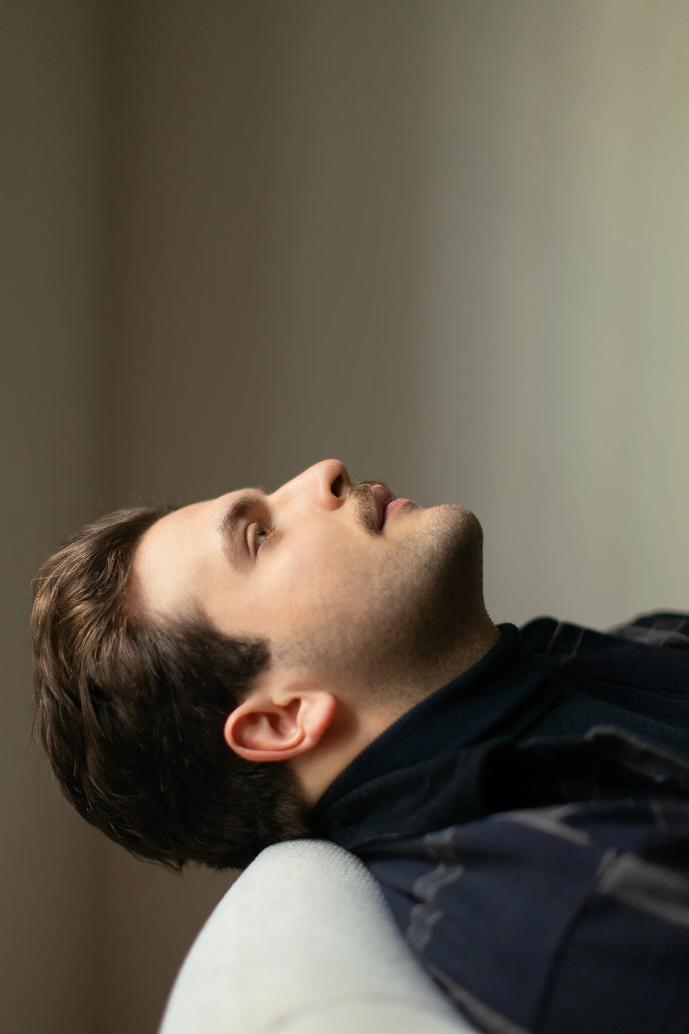 Un homme allongé, vu de profil, reposant sur un coussin blanc, regardant vers le haut, avec un fond simple.