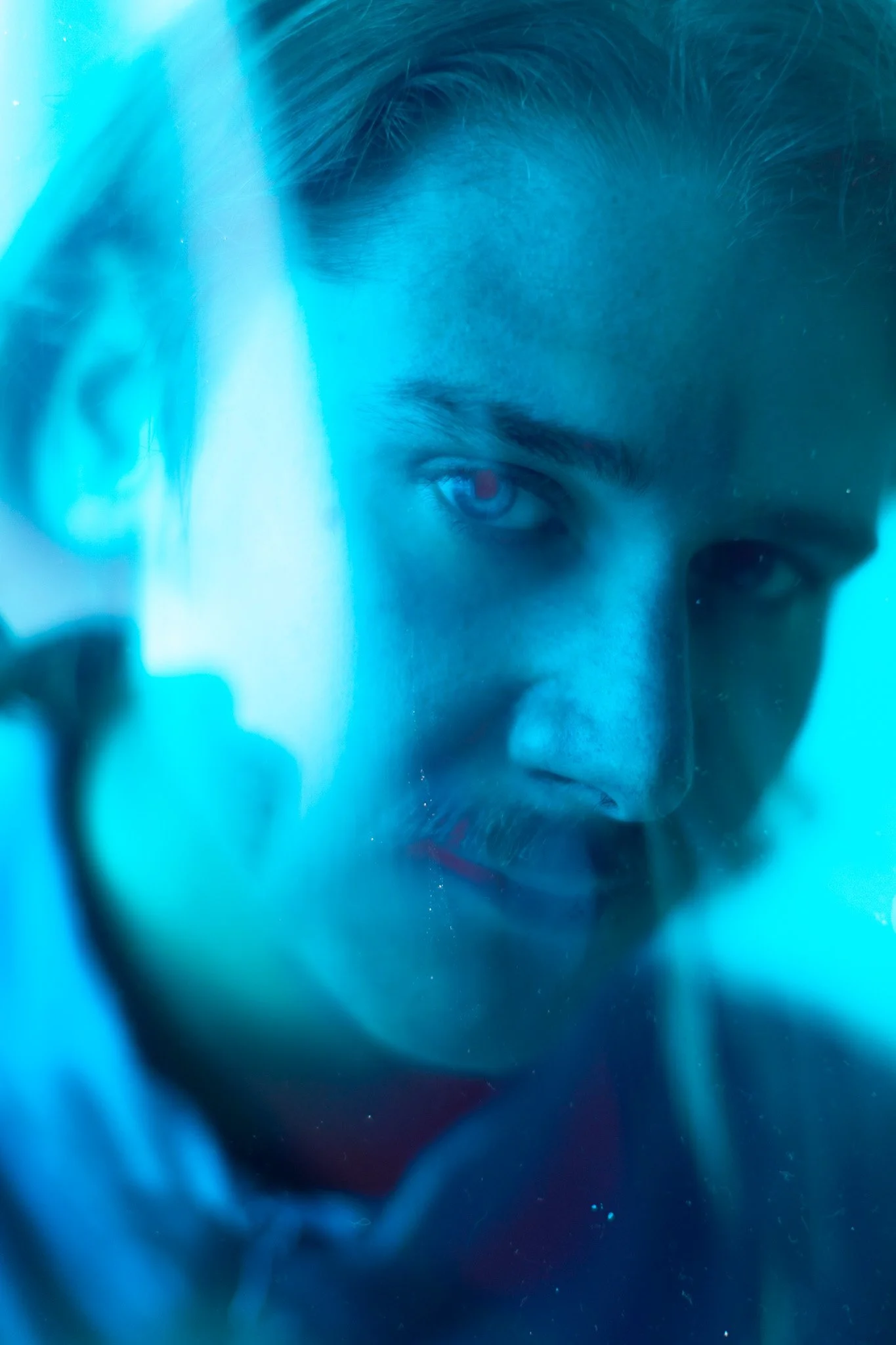 Gros plan d'un homme avec des cheveux foncés, regardant intensément, avec un éclairage bleu qui crée une ambiance mystérieuse.