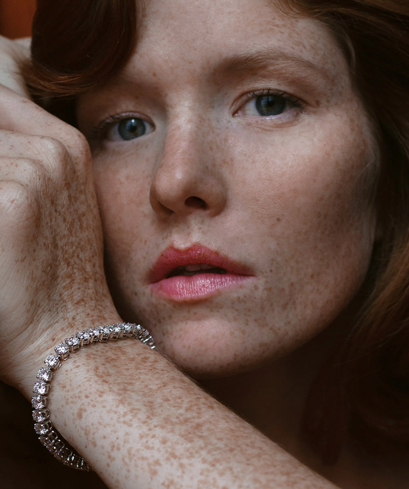 Un gros plan d'une femme aux cheveux roux, à la peau claire et parsemée de taches de rousseur, portant un bracelet en diamants, avec une expression neutre.