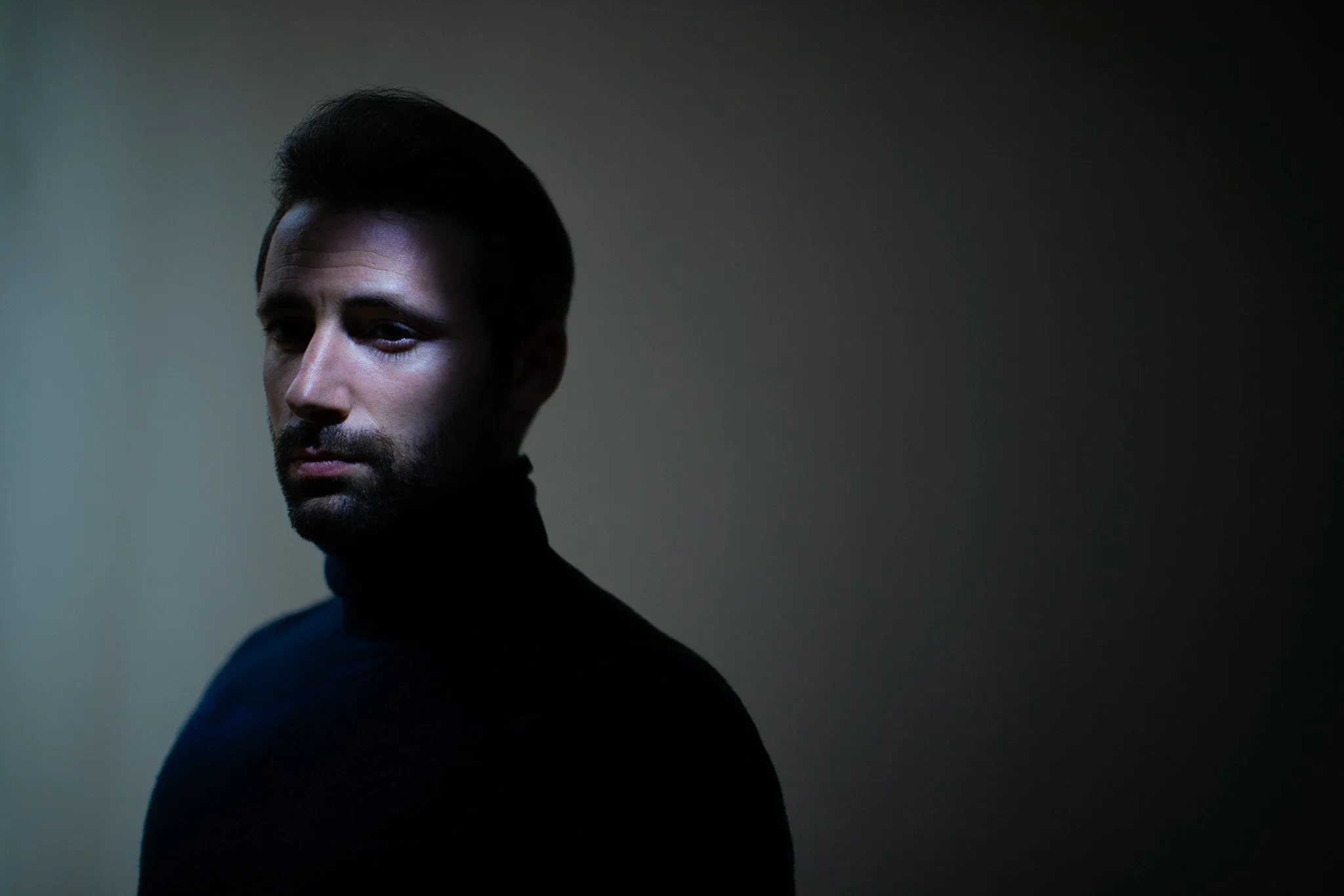 Un homme avec une barbe porte un pull noir, regardant vers la gauche, dans une lumière tamisée.