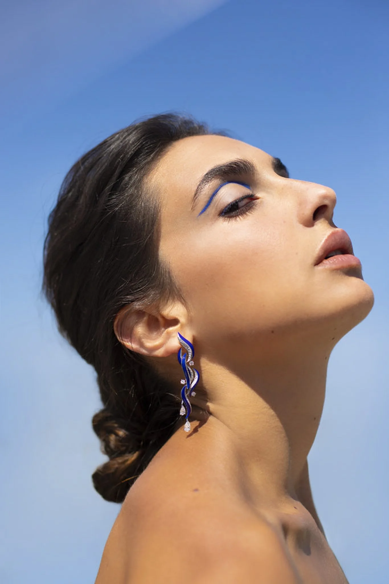 Une femme avec une boucle d'oreille élégante, portant du maquillage coloré, pose contre un ciel bleu.