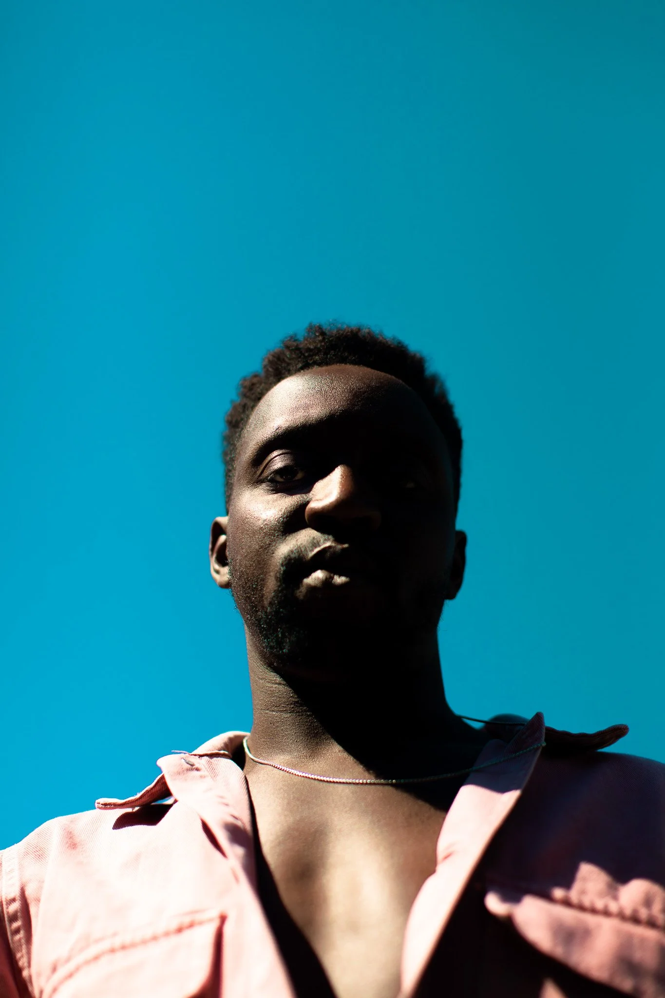 Portrait d'un homme noir avec un haut rose, prise en contre-plongée avec un ciel bleu en arrière-plan.