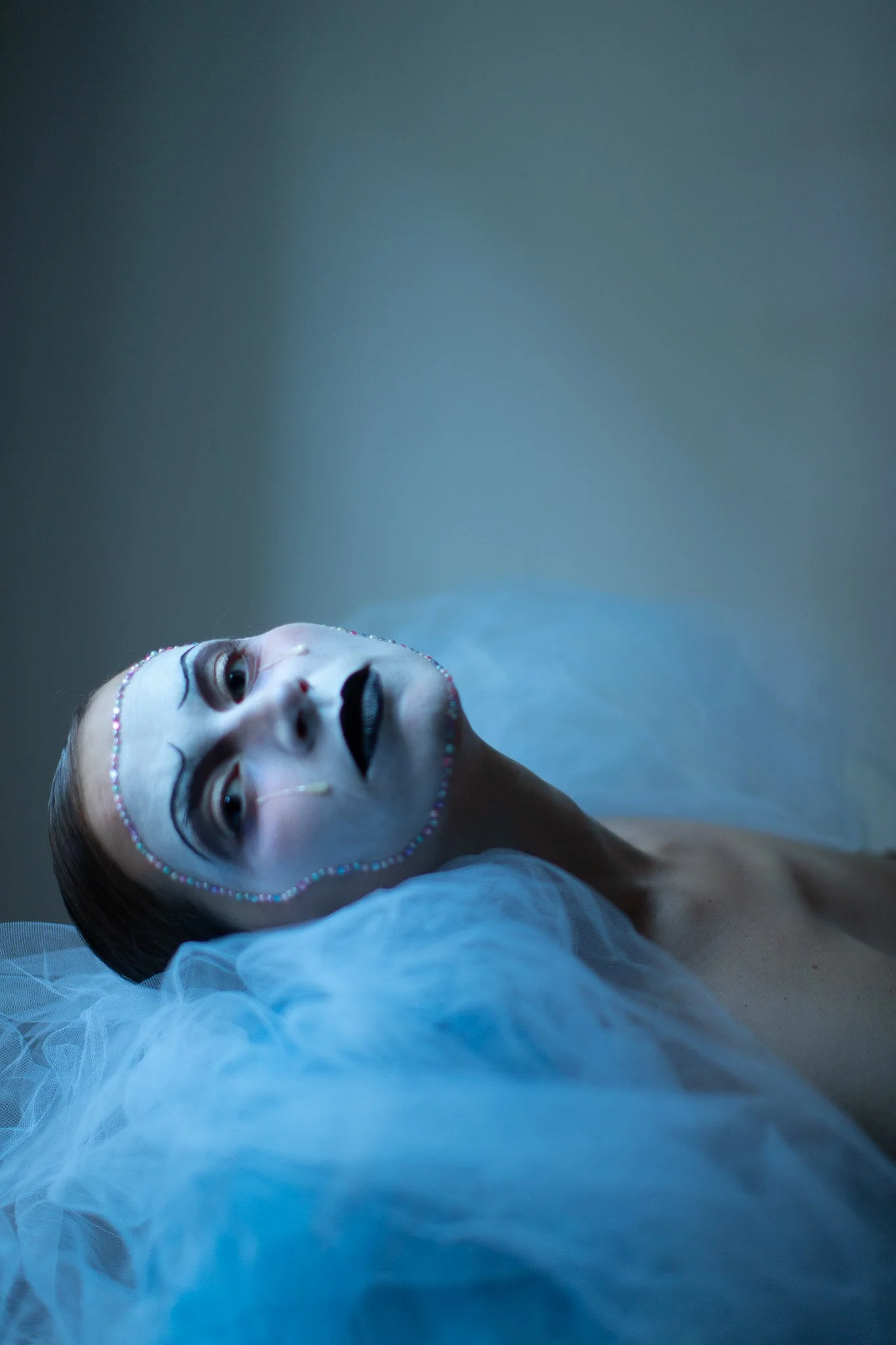 Portrait d'une personne féminine avec maquillage artistique et décorations de perles sur le visage, allongée sur un tissu blanc, éclairée par une lumière douce et froide.