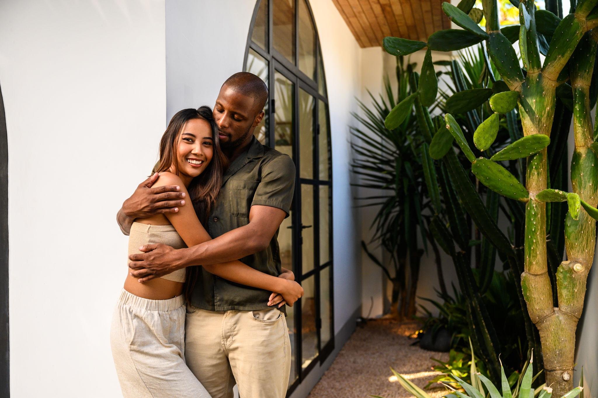 A young woman and a man hugging, standing outside a modern building with large windows and surrounded by tall green cacti and plants.