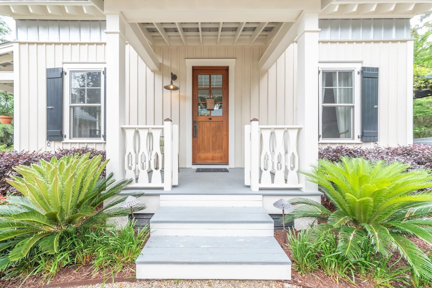 Front porch of a white house with a brown wooden door, surrounded by green plants and shrubbery, with two small steps leading up to the porch.