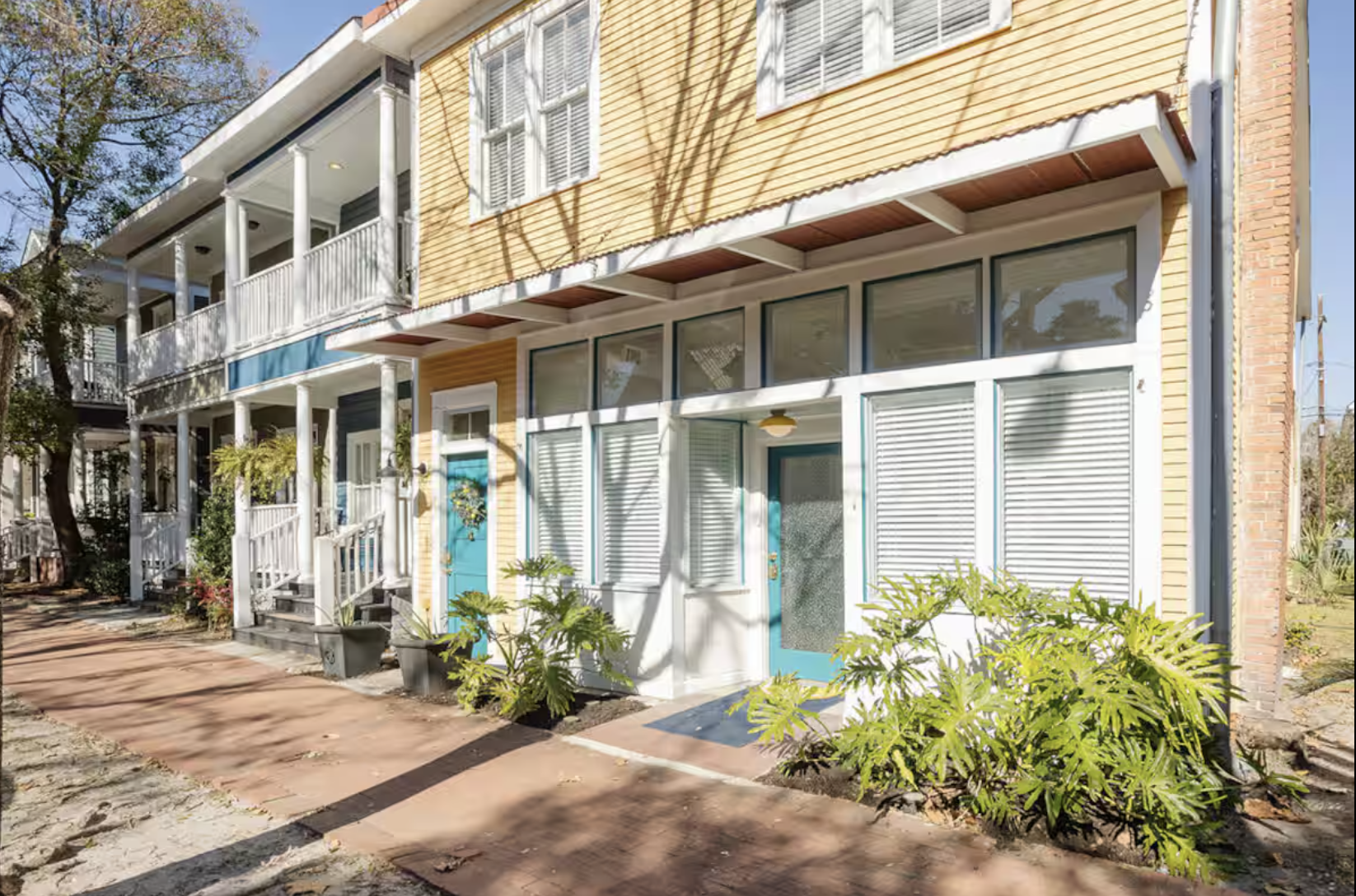 A colorful multi-story residential building with yellow, white, and gray siding, featuring balconies and a front door with potted plants and sidewalk in front.