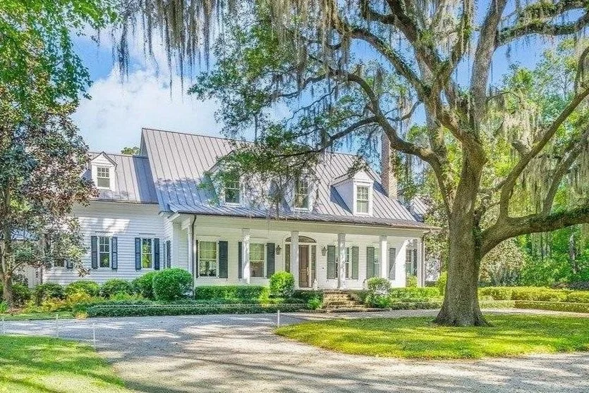 Large white house with a metal roof, front porch with columns, surrounded by lush green trees and manicured lawn.