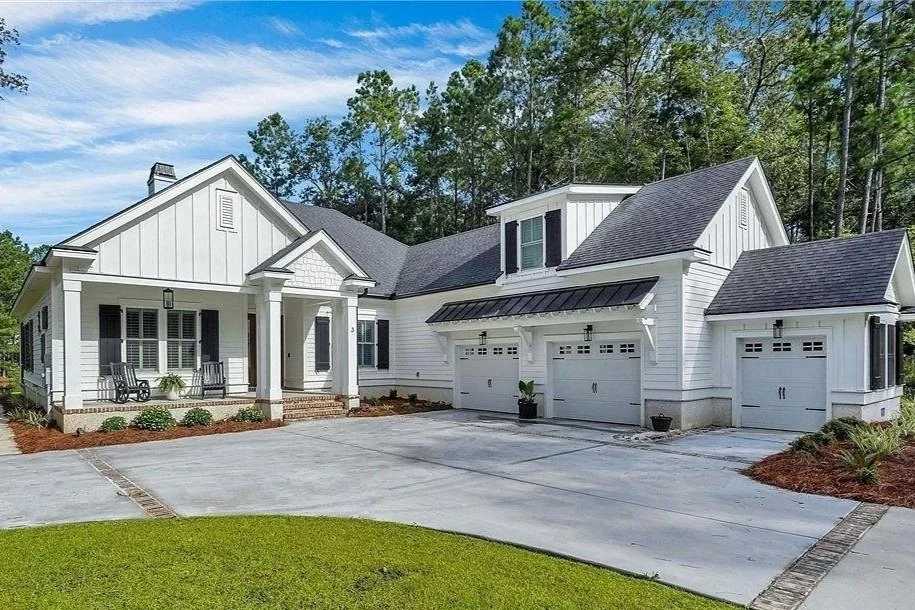 White modern house with multiple garages, a front porch with chairs, and a well-maintained lawn, surrounded by trees.