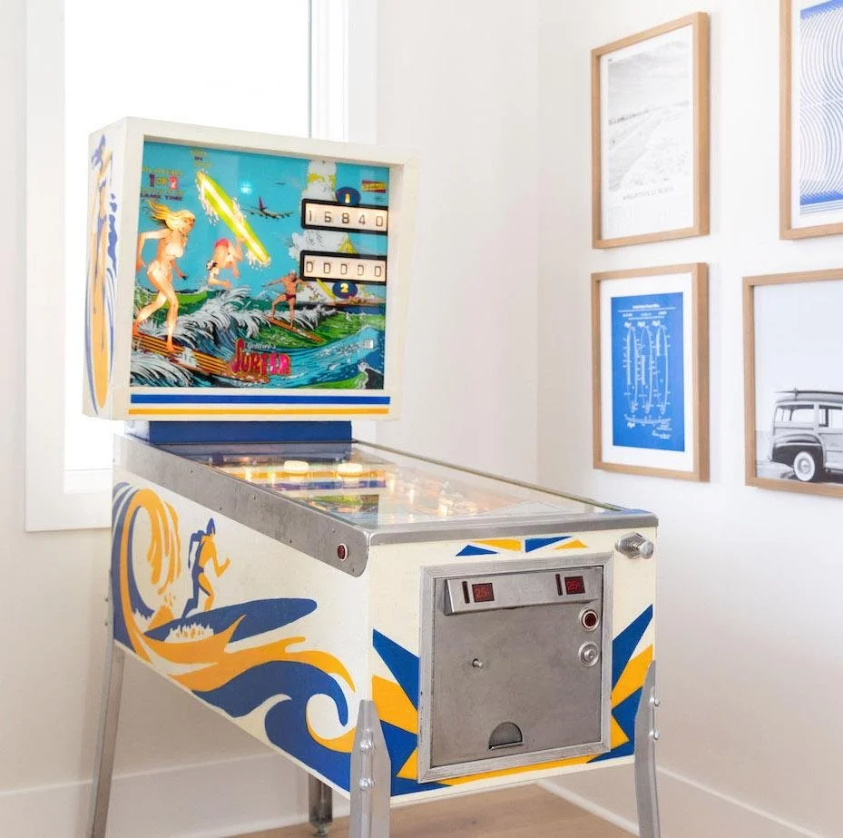 A vintage Surf Champ pinball machine with a beach and surfing theme, featuring a digital score display, located in a room with framed artwork on the wall.
